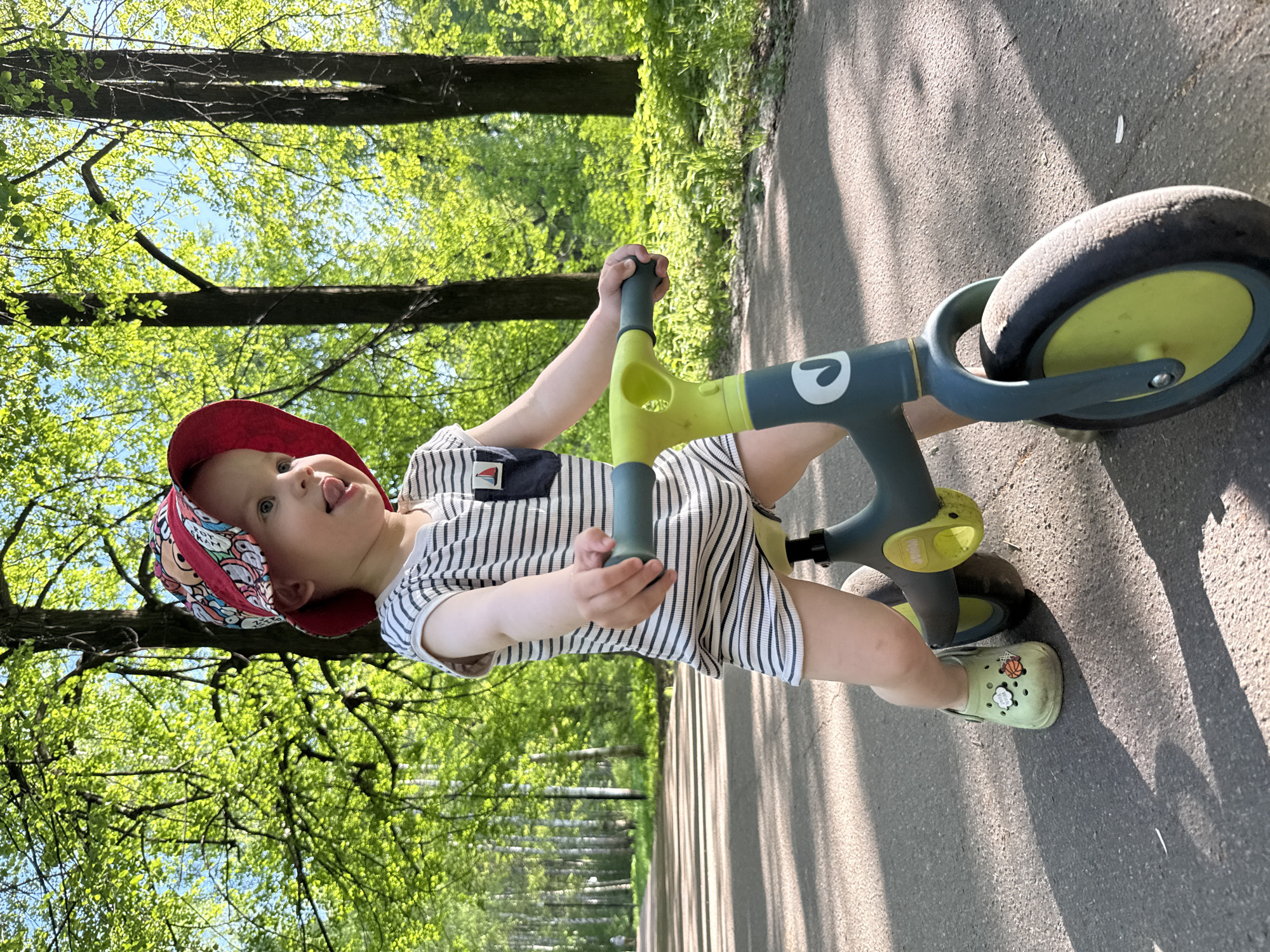 A young child is standing on a gray paved path, holding onto the handlebars of a child's balance bike. The child is wearing a red hat with a colorful pattern, a white and black striped shirt, and light-colored shoes. The balance bike has a green frame and gray wheels. The background shows a park with green trees and a glimpse of blue sky. The photo appears to be taken outdoors in a park-like setting.