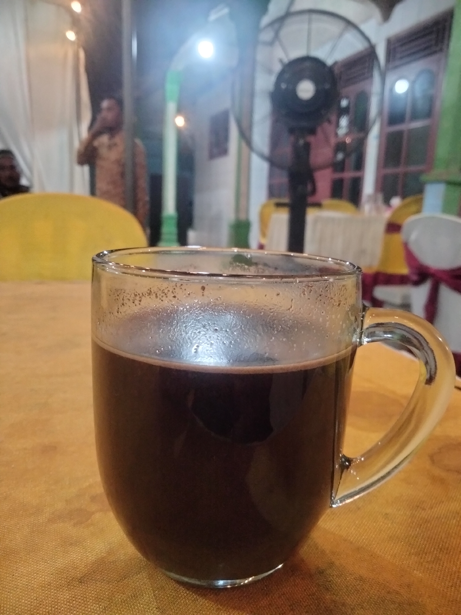 The image shows a close-up of a clear glass mug filled with a dark liquid, likely coffee, resting on a textured orange surface. The mug has condensation on the outside. In the blurred background, several elements suggest an indoor setting with some elements of an outdoor or semi-outdoor space. A person in a patterned shirt is visible in the middle ground, appearing to hold a cup to their mouth. To the right, a large black standing fan is prominent, with its blades blurred, indicating it may be in operation. Behind the fan, windows with dark frames and panes are visible, suggesting a building. There are also hints of yellow and white fabric draped over furniture, possibly chairs or tables, and some lighting fixtures can be seen, contributing to a dimly lit atmosphere. The overall impression is of a casual gathering or a cafe setting. The reference to Ketanggung, Indonesia, suggests this scene is located in that specific geographical area.