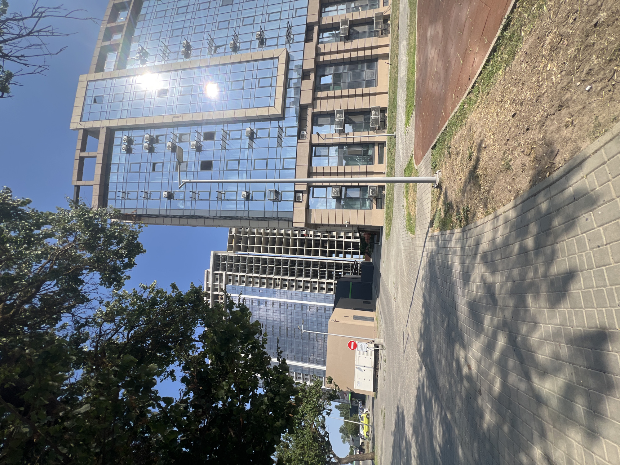 The image depicts a street scene in Fontanka, Ukraine, on a clear, sunny day. In the foreground, a paved walkway with diamond-shaped patterns is visible, casting long shadows from an unseen tree on the left. Adjacent to the walkway is an area of dry, patchy grass and exposed earth, bordered by a red-colored rubberized surface.
Dominating the background are several modern buildings. On the right, a multi-story building with a glass facade and beige stone accents stands prominently. Numerous air conditioning units are mounted on the exterior of this building, and the sun reflects brightly off its windows. To the left of this building, a much taller, unfinished residential complex is under construction, its concrete framework exposed. Further to the left, another completed residential tower with a reflective glass exterior is visible.
In the middle ground, a small, beige, rectangular building with dark green accents is situated. A shipping container is parked in front of it, and a red and white circular "no entry" sign is mounted on a pole. A light pole with multiple fixtures stands in front of the glass-fronted building. A bright yellow car is visible in the distance to the left of the beige building. The sky is a clear, vibrant blue.