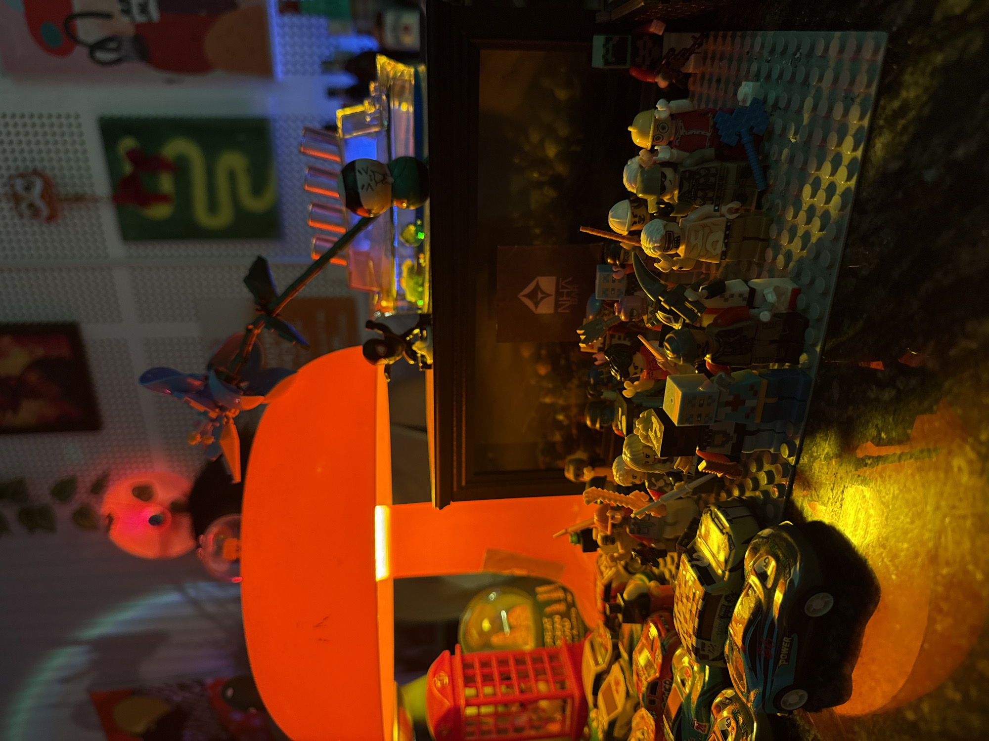 The image captures a vibrant tabletop display of toys, primarily LEGO, illuminated by various artificial lights in a low-light setting. The scene is likely indoors, at night or late evening, creating a warm and dramatic ambiance.
The main subjects are a large assembly of LEGO minifigures standing on a grey studded baseplate. These minifigures appear to be a diverse collection, some armed with miniature weapons like swords, axes, and firearms, suggesting a battle or adventure scenario. Behind them, several LEGO structures resembling city buildings or light fixtures glow with blue and yellow lights.
To the right, several toy vehicles are visible, including a dark blue sports car with the word "POWER" written on its side, and a miniature red double-decker bus. A large, bright orange/red circular lamp dominates the left side of the frame, casting a strong, warm glow over the immediate foreground and creating distinct shadows. Above this lamp, a blue toy with a propeller-like element is visible.
In the blurred background, a textured wall (possibly a pegboard) displays framed artworks, including a green rectangular piece with a yellow wavy line and a red piece showing part of a cartoon character. A glowing pinkish object resembling a skull or stylized face is also in the background. A dark rectangular frame in the mid-ground contains a small black sign with a white logo (a stylized upward arrow/triangle) and the text "NHX". The overall setup appears to be a carefully arranged display or a miniature world for imaginative play. The location context for this scene is Đà Nẵng, Vietnam.