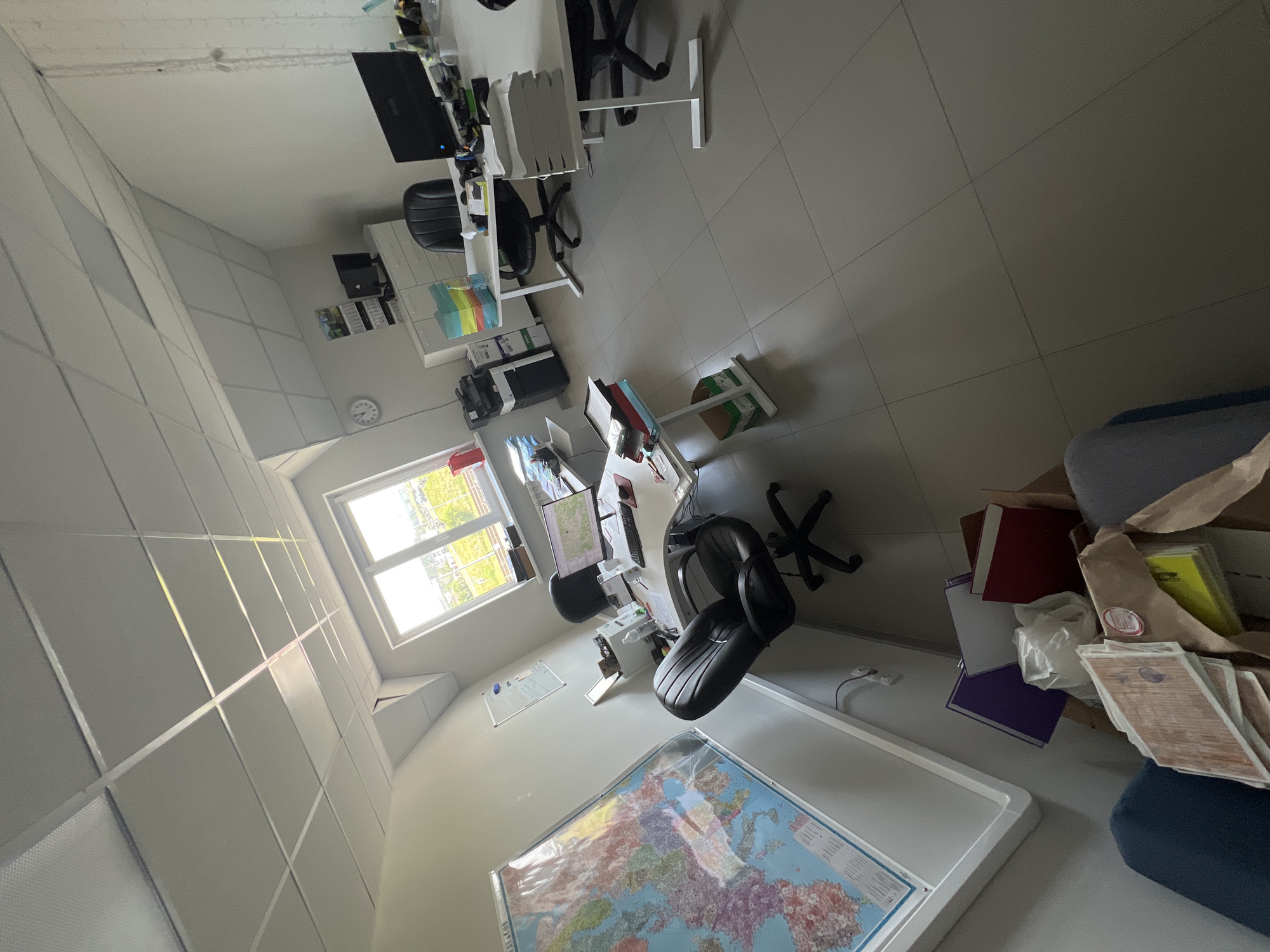 The image depicts an office space with multiple desks and chairs. On the left, a large map of Europe is displayed on the wall. Several stacks of paper are visible on the desks, along with computer monitors and printers. Two black office chairs are positioned at desks, and another black office chair is in the foreground. The room has a tiled floor and a suspended ceiling with fluorescent lighting. A window on the right side of the room offers a view of an outdoor area with greenery. In the lower right corner, a box contains stacks of papers and folders. The overall impression is that of a functional, albeit somewhat cluttered, office environment. The image does not provide any specific clues that would allow for localization to Ждановіцкі сельскі Савет, Belarus.