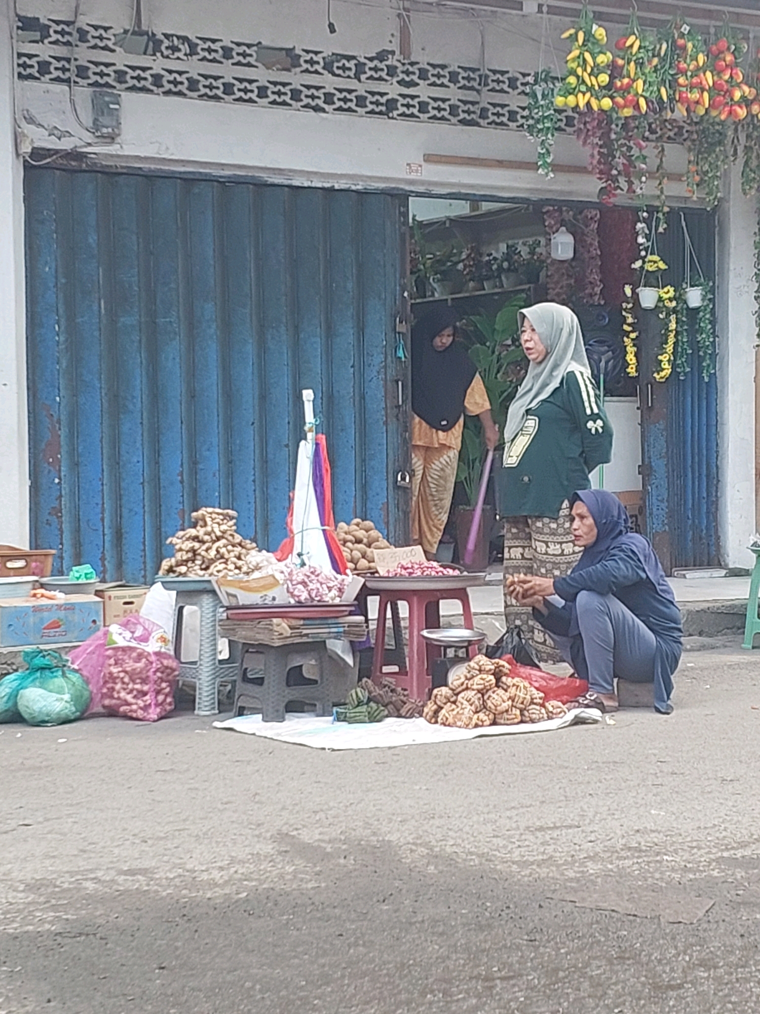 A street scene in Ambon, Indonesia, features a small shop with a blue, corrugated metal roll-up door partially open, revealing shelves stocked with plants and decorative items. In front of the shop, on the paved street, a vendor is selling produce. Three women are visible. One woman, wearing a black hijab and an orange and yellow patterned garment, stands in the doorway. Another woman, wearing a light grey hijab and a green shirt with an abstract design, stands near the entrance, facing outwards. A third woman, in a dark blue hijab and blue pants, crouches on the ground to the right, attending to a pile of goods.
In the foreground, various food items are displayed on small stools and tables. These include large piles of ginger, onions in mesh bags, and other root vegetables. A red table holds a pile of pink flowers and a scale. Another stool has a stack of what appear to be papers or books. The ground is asphalt, and the overall lighting suggests it is daytime. Decorative hanging plants and flowers adorn the upper portion of the shopfront.
