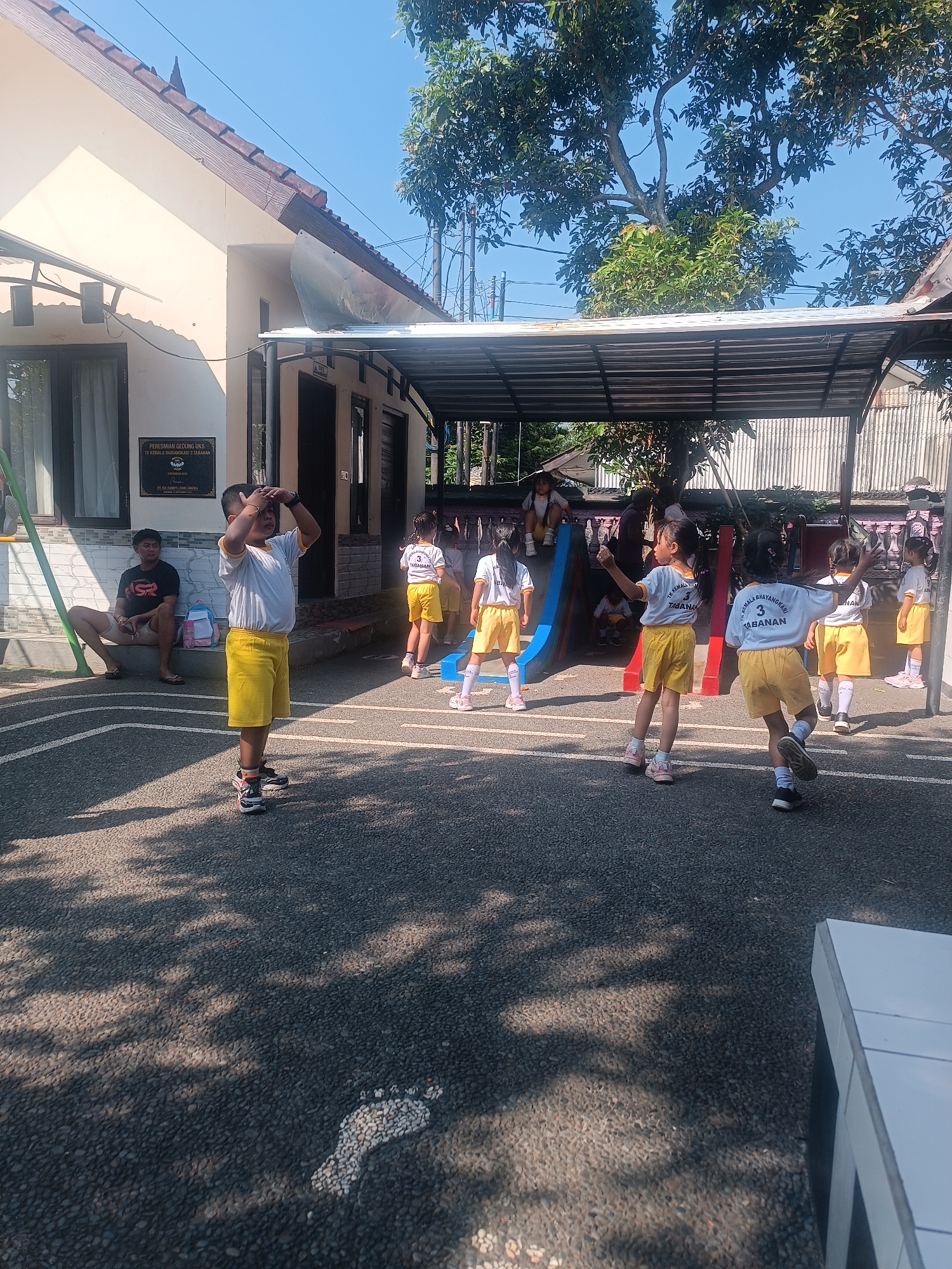 In an outdoor setting, likely a schoolyard in Tabanan, Indonesia, several young children are engaged in an activity. The children are wearing matching white t-shirts with yellow collars and yellow shorts, suggesting a school uniform. Some of the children are facing away from the camera, appearing to be listening to instructions or observing something in front of them. One boy in the foreground is standing and has his hands on his forehead. In the background, there is a building with a tiled roof and a covered outdoor area with a metal roof. A man is seated on a step near the building, observing the children. The ground is paved with asphalt and there are painted lines on it, possibly indicating a playing field or track. Trees and foliage are visible in the background, indicating a sunny day.