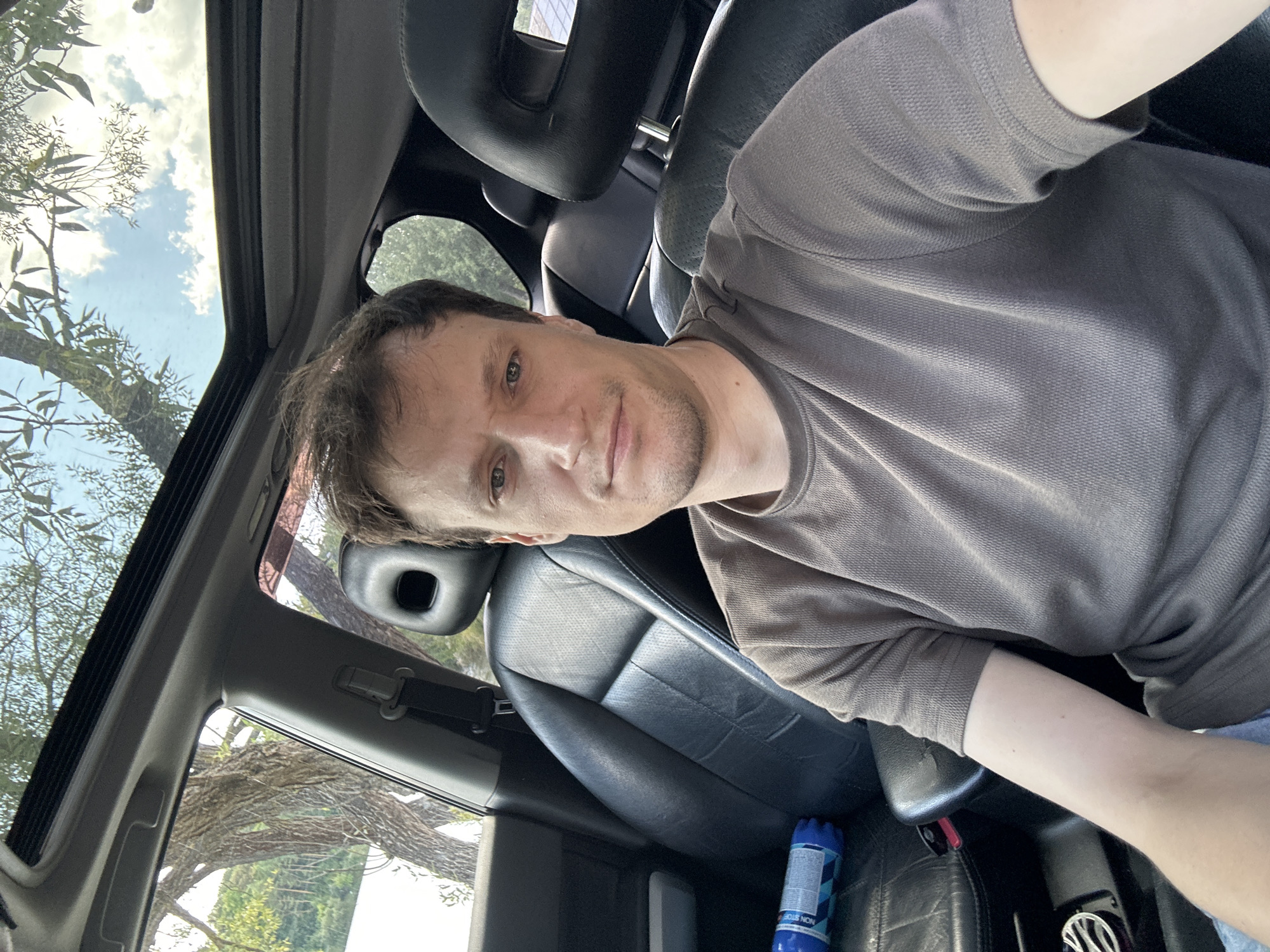 A man is seated in the driver's seat of a car, taking a selfie. He is wearing a grey t-shirt and has short brown hair. The interior of the car is visible, including black leather seats and a sunroof which is open, revealing a tree and sky. A blue water bottle is on the passenger seat. The car appears to be parked outdoors, possibly near a body of water, judging by the reflection of trees and sky. The scene is illuminated by natural daylight. The image was taken in Makiv, Ukraine.