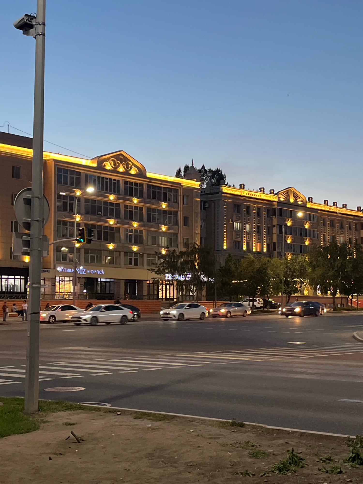 The image depicts a street scene at dusk in Astana, Kazakhstan. A large, multi-story building with illuminated architectural details and windows lines the right side of the frame. The building appears to be a mix of commercial and residential spaces, with signage indicating a business named "Optika Koz" on the lower level. Several cars are visible on the road in front of the building, with some moving and others stopped at a pedestrian crossing. A traffic light shows a green signal. A lamppost with a traffic camera is positioned on the left side of the image, near the foreground. Trees line the sidewalk in front of the building, and the sky is a clear twilight blue. A few pedestrians are seated on a bench near the building. The overall scene conveys an urban environment in the early evening.
