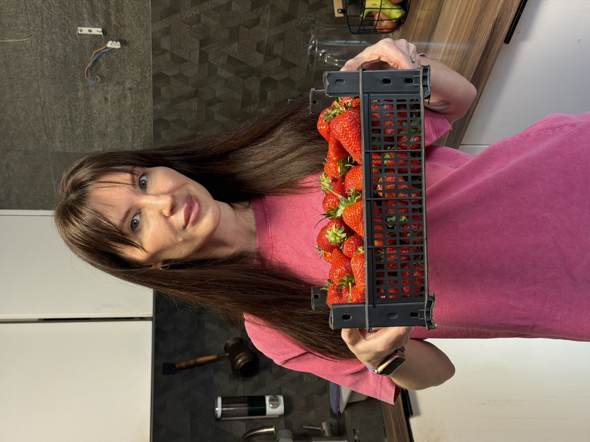 A woman is holding a crate of strawberries in what appears to be a kitchen. The kitchen has dark gray tile walls, and a light-colored countertop. She is wearing a pink shirt. The image was likely taken in Perm, Russia.