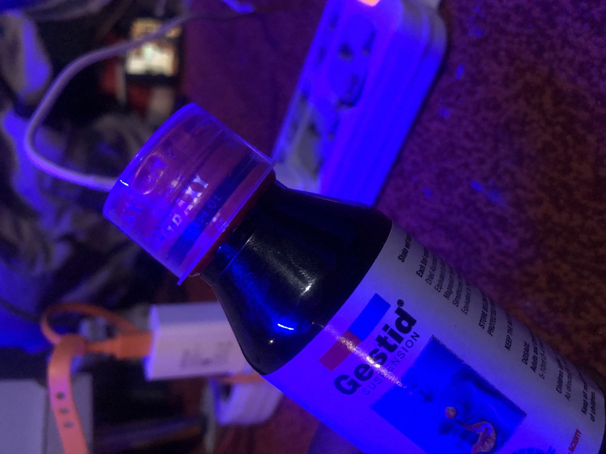 The scene is dominated by a strong blue light source, casting a blue-purple hue across all visible objects and surfaces. In the foreground, a dark-colored plastic bottle with a white label is angled towards the viewer. The label prominently displays "Gestid" with a registered trademark symbol, followed by "SUSPENSION". A red and blue horizontal stripe design is visible on the label. Partial text on the label includes "Shake well before...", "Each 5ml cont...", "Dried Alumin...", "Magnesium Hy...", "Simethicone...", "STORE BELOW...", "DOSAGE", "Adults and Children", and "Children". The bottle is topped with a clear plastic cap, appearing to be a measuring cup, which reflects the blue light with a purple tint. Text visible on the cap includes "DAXY" and what appears to be "10 ML".
The bottle rests on a textured surface, likely a dark red or purple fabric with a coarse, patterned weave, which also reflects the blue light. To the right of the bottle, in the mid-ground, a white power strip (surge protector) with multiple outlets is visible, illuminated by the blue light. A small orange indicator light is visible on its far right end. A white cable runs from behind the bottle towards the power strip.
To the left of the bottle, in the mid-ground, a white wall charger block is present. An orange cable is plugged into this charger, curving upwards and forming a loop before exiting the frame to the left. Behind the charger, partially obscured, is an object with small, circular perforations, possibly a watch band. The background consists of indistinct, dark shapes and blurry areas, further illuminated by the blue light, suggesting other objects or furniture, with another white cable snaking through. No people are visible in the image. The location is in the city of Unknown, Unknown.