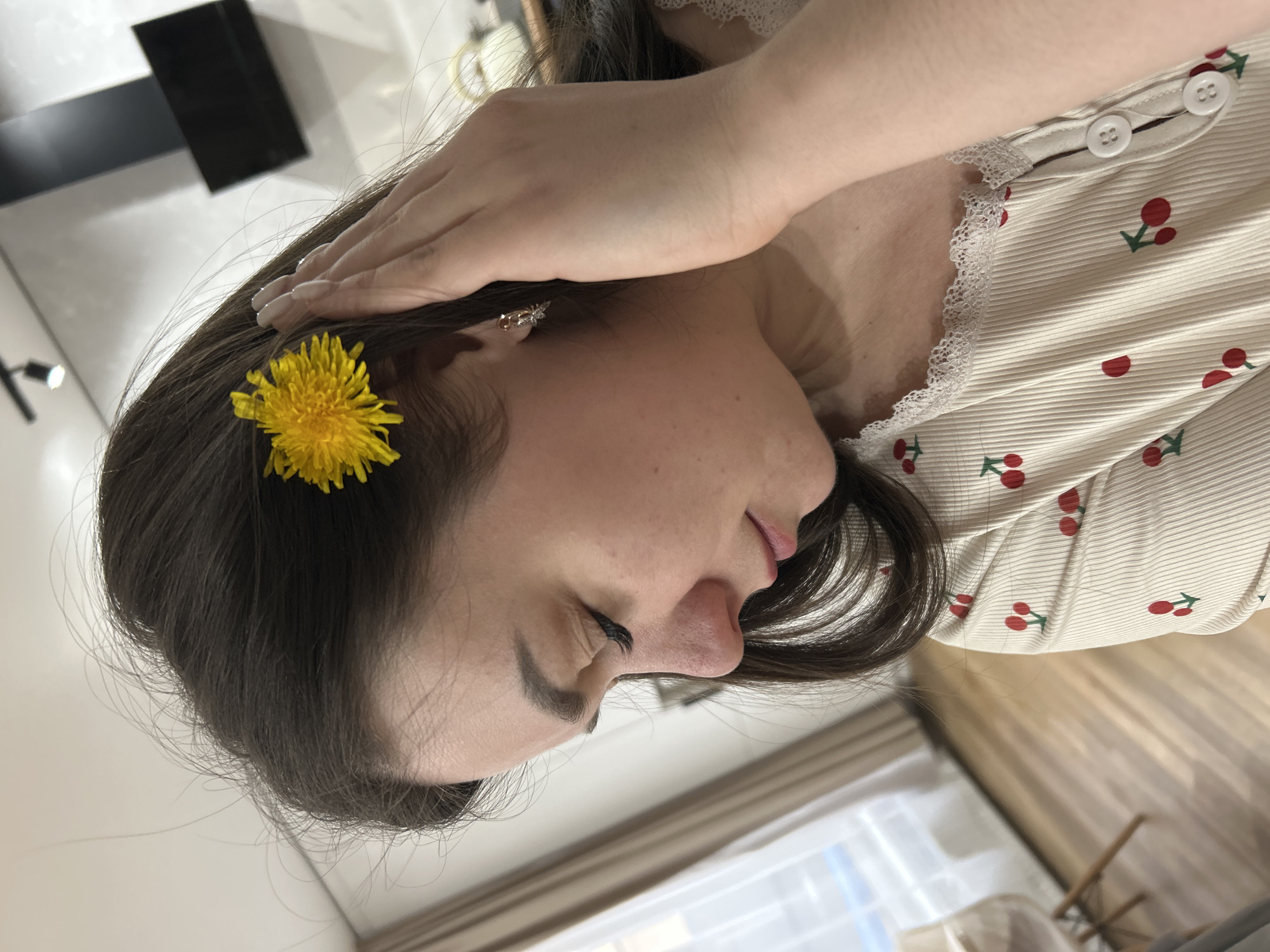 A woman with dark hair poses indoors. A yellow dandelion flower is tucked into her hair near her left ear, adorned with an earring. Her hand rests near her forehead. She wears a cream-colored top with red cherry prints and white lace trim. The location is implied to be Chaikovsky, Russia, although this cannot be determined from the image alone. The background features a window, parts of furniture, and a black rectangular object.