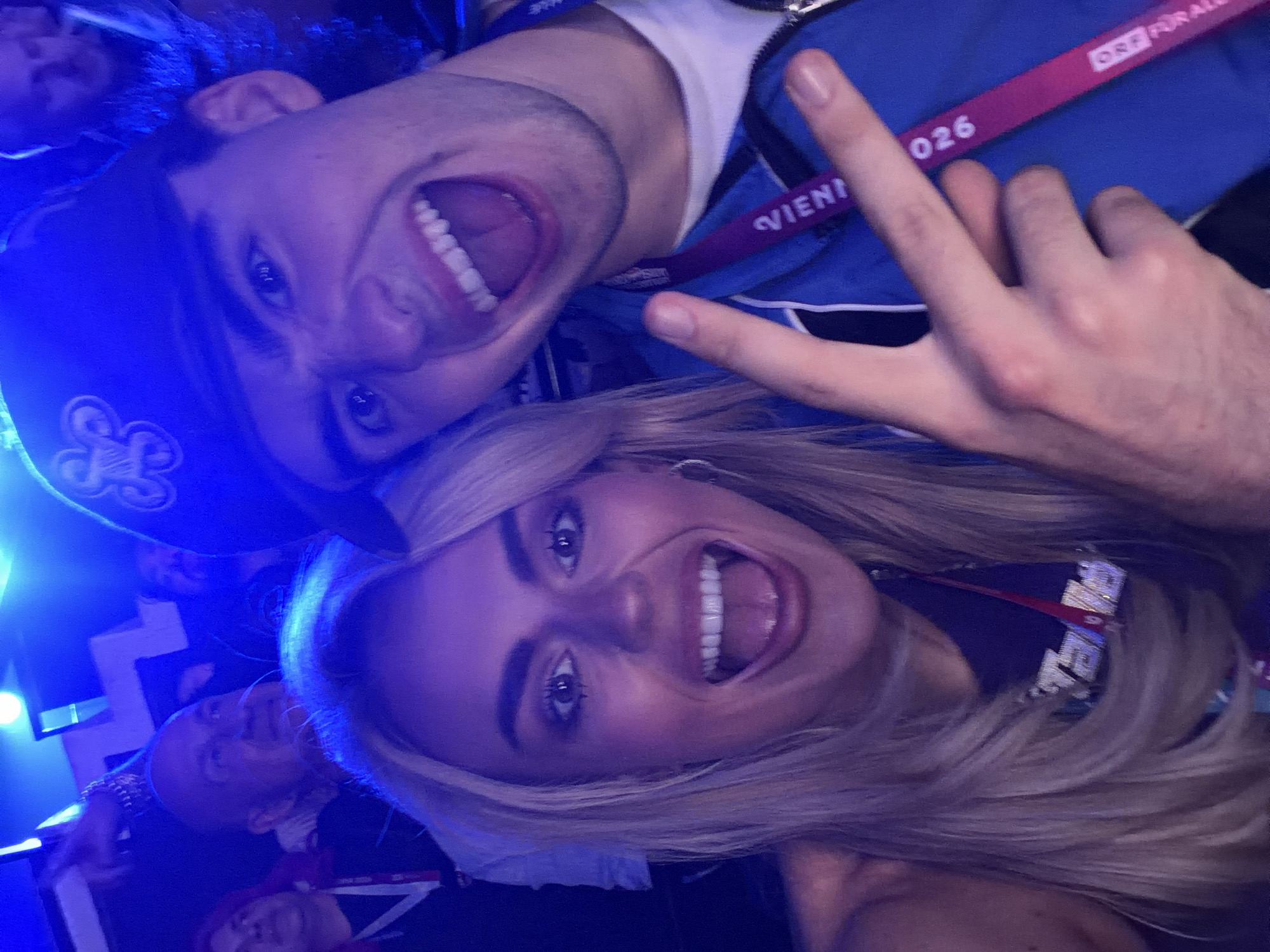A close-up, selfie-style photograph shows two people, a man and a woman, smiling widely with their mouths open. They are positioned close together. The man is wearing a blue hat with a design on it and a blue jacket with a lanyard around his neck that reads "Vienna 026." He is making a peace sign with his fingers. The woman has long blonde hair and is also wearing a lanyard. In the background, there are other people and bright blue lighting. Based on the lanyard and context clues, the location is likely Vienna, Austria, where the lanyard seems to have been issued. The photograph appears to have been taken at an event or gathering.
