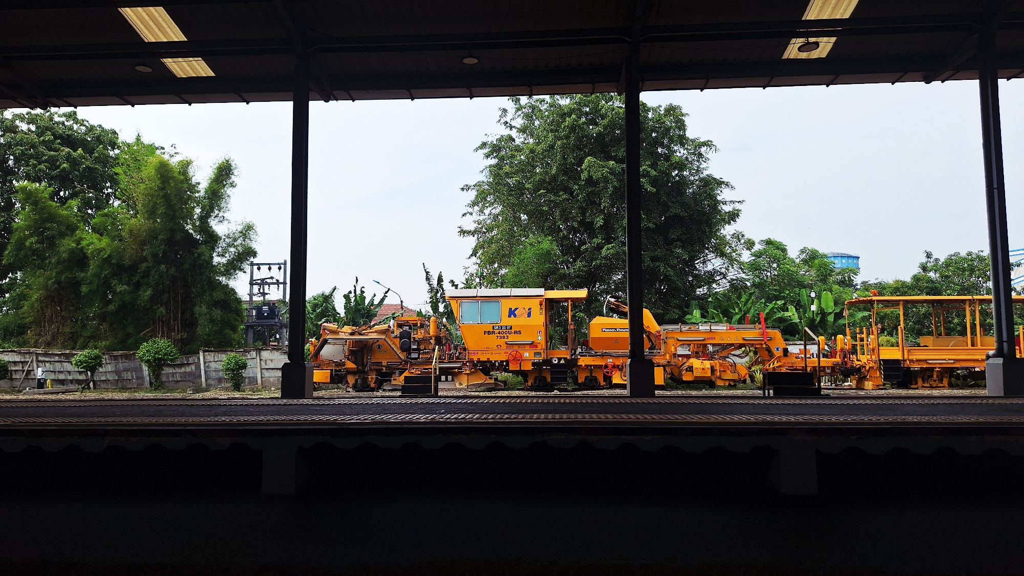 The image shows a railway platform view, likely in Karawang, Indonesia, showcasing specialized railway maintenance vehicles. The machinery, colored in yellow and orange, appears to be for track tamping and adjustment. The vehicles sit on rails next to a raised platform. Behind the equipment, trees and vegetation form a backdrop with a pale sky. A building structure frames the scene.