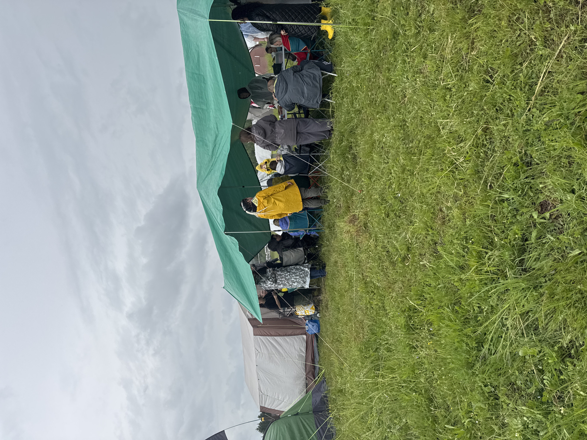 This outdoor scene, set in Bol'shaya Kivara, Russia, captures a group of people gathered under a large green tarp shelter on an overcast day. The sky, filled with dense clouds, occupies the upper portion of the image, while a sprawling grassy field dominates the right side and foreground. Several individuals are visible, mostly seated on camping chairs beneath the green tarp, which extends horizontally across the frame. They are dressed in warm, casual attire, with some wearing raincoats, including a person in a prominent yellow jacket in the left foreground.
The gathering suggests a camping trip or an outdoor event, with the tarp providing shelter from what appears to be cool or potentially rainy weather. In the background, to the left of the group, a large brown and white tent is pitched, with a smaller green and black tent partially visible further left. Guylines from the tents and tarp stretch across the grassy ground. Camping gear, such as a blue container, is seen near the tents. The grass in the field is long and natural, indicating an uncultivated outdoor environment. No discernible text is visible in the image.