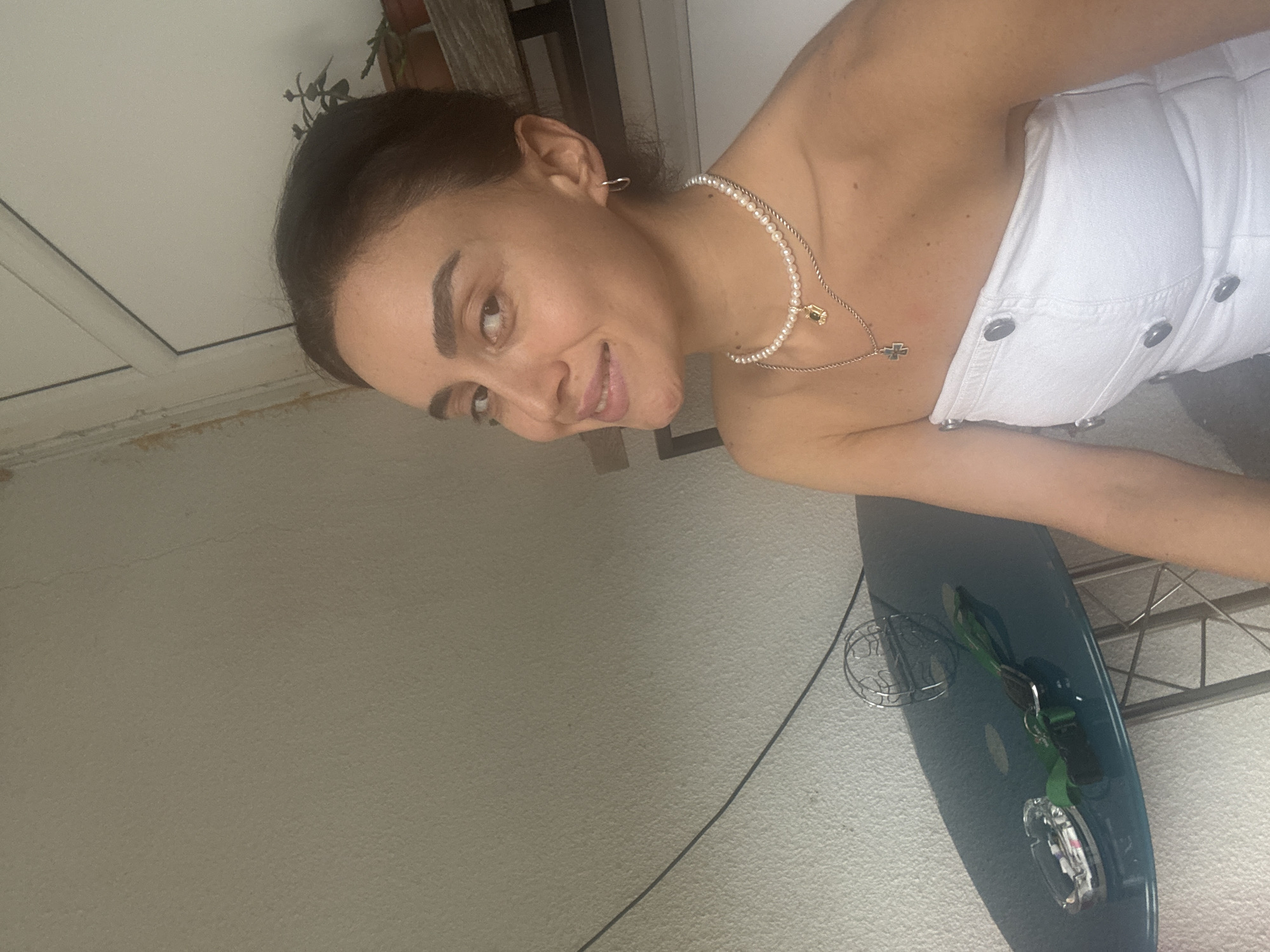 A woman is taking a selfie on a balcony. She is wearing a white strapless top and a pearl necklace. She has dark hair pulled back and is smiling at the camera. On a small glass-topped table next to her is an ashtray with cigarette butts and a green leash. The background is a plain wall and a railing. The scene suggests a residential area, possibly in Tbilisi, Georgia, given the presence of plants on a shelf.