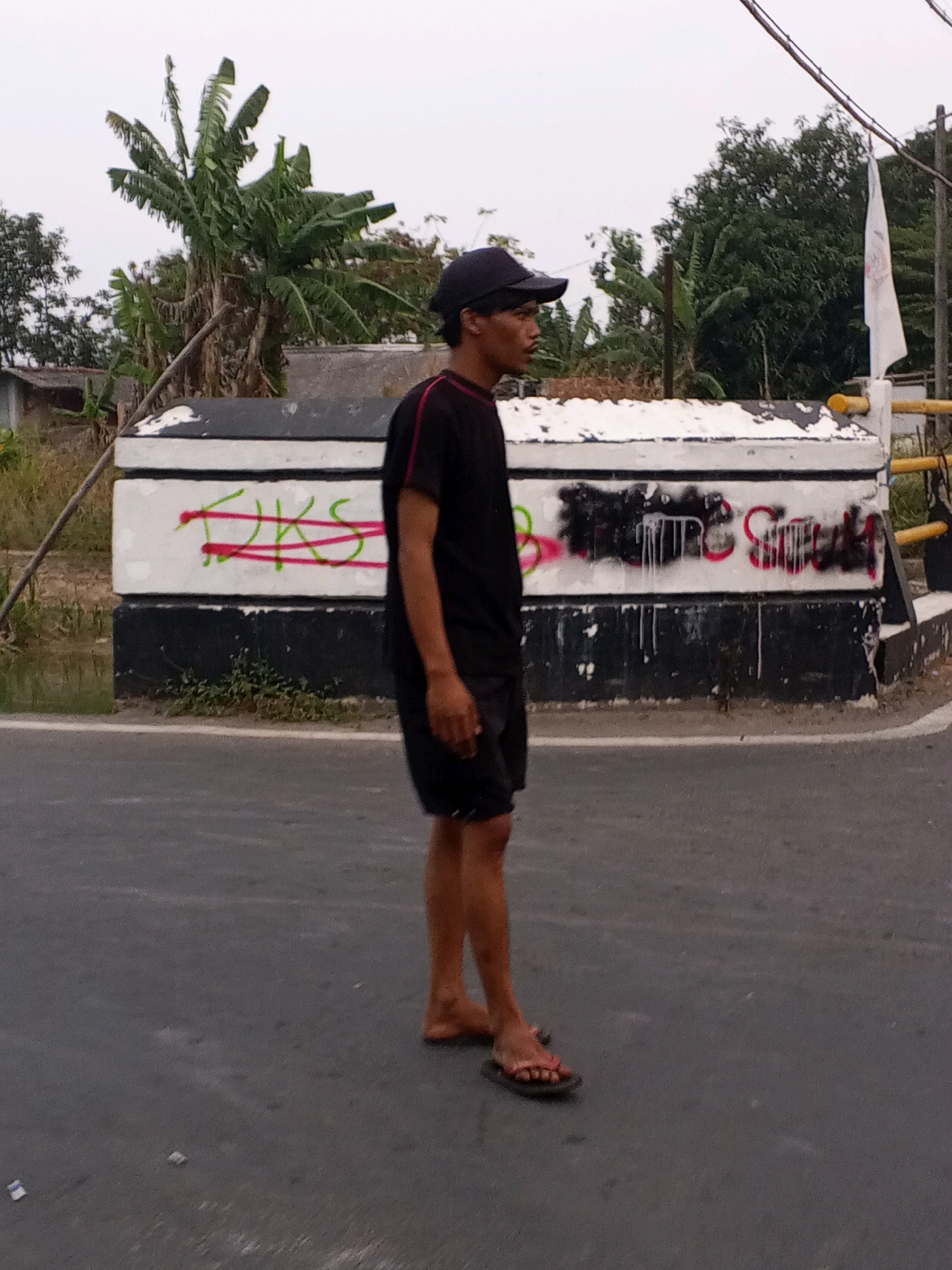 In the city of Ciruas, Indonesia, a young man wearing a black t-shirt, black shorts, and a black baseball cap stands on an asphalt road, facing to the right. He is barefoot, wearing only flip-flops. Behind him is a low, concrete structure covered in white paint and graffiti. The graffiti consists of red and green markings on the upper section and black spray paint with white drips on the lower section. To the left of the man is a lush green banana tree. In the background, more trees and a white flag on a pole are visible. The sky is overcast.