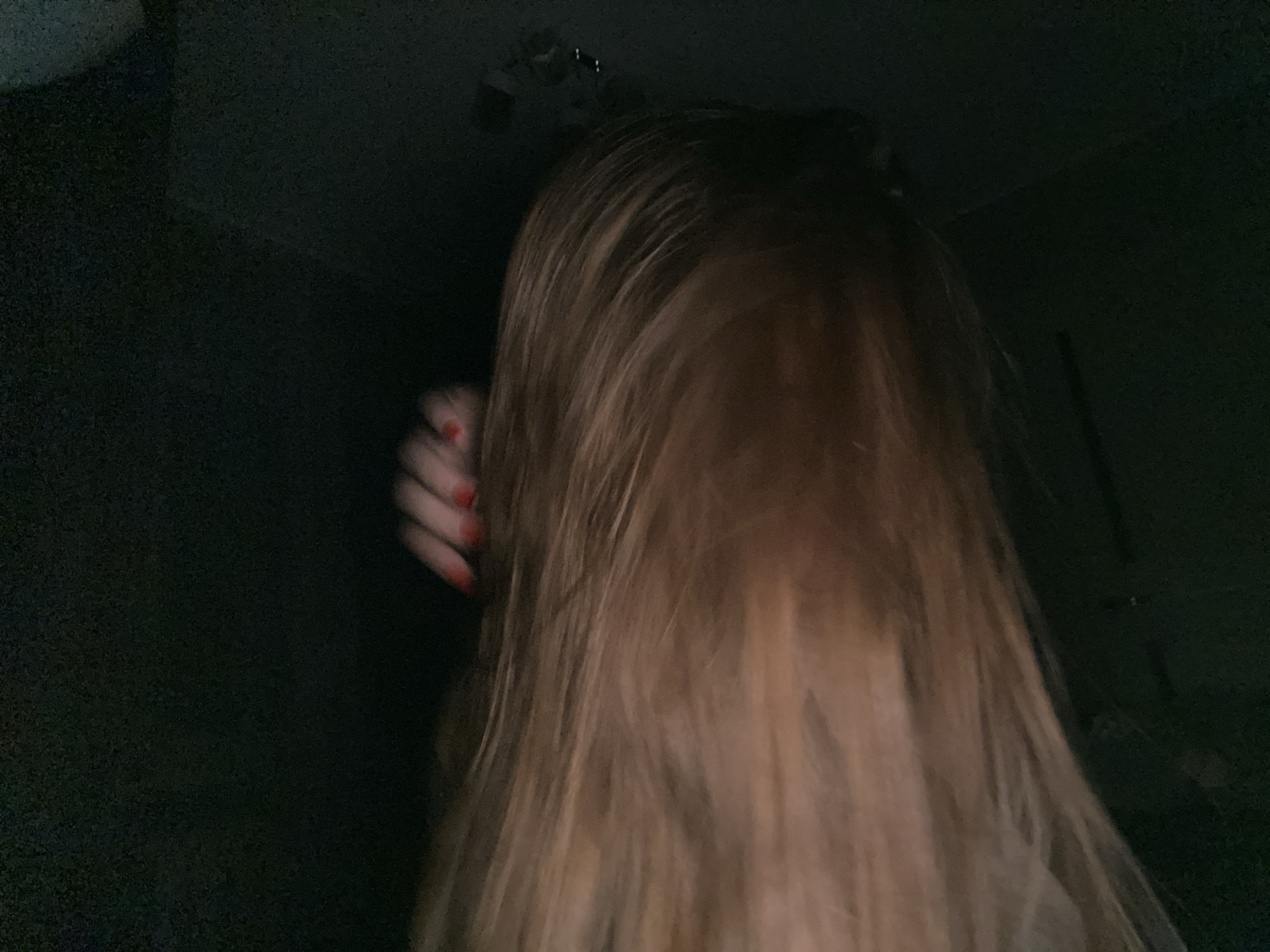 The image, taken in a dimly lit indoor environment, focuses on the back of a person's head, with their long, light brown hair obscuring most of their face. A hand with red nail polish is visible on the left side of the frame, positioned near the person's hair. The background is dark and indistinct, offering no clear clues about the specific location within Dyakovo, Russia. The overall impression is one of low light and a close-up perspective.