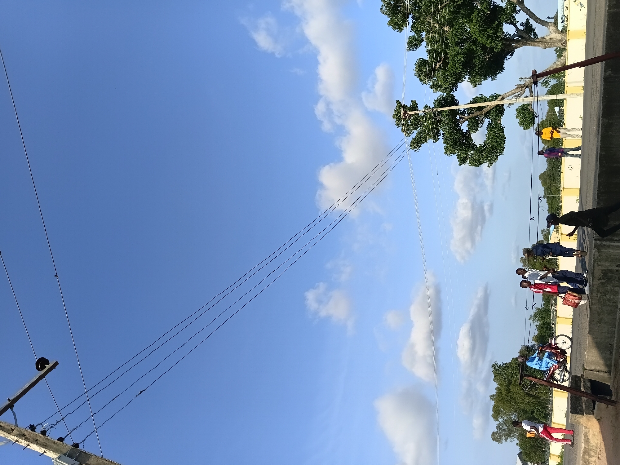 This is a full-shot photograph of a street scene in Jalingo, Nigeria, with a clear blue sky and scattered white clouds. Overhead, power lines stretch across the frame, supported by a wooden pole on the left and another pole with a large tree on the right. On the right side of the road, a man in a yellow shirt and light-colored pants stands near the pole. To his left, two young men, one in a purple shirt and the other in a red and white checkered shirt, stand together. Further left, a young man in a white shirt and dark pants, holding a red and white bag, walks. A motorcycle, ridden by a man in a blue outfit, is in motion on the road. On the far left, a person in a white shirt and red pants walks away from the camera. The road is bordered by a concrete structure on the bottom and a yellow wall in the background. Trees and vegetation are visible behind the wall. The overall impression is of a bright, possibly warm, day in an urban or semi-urban environment.