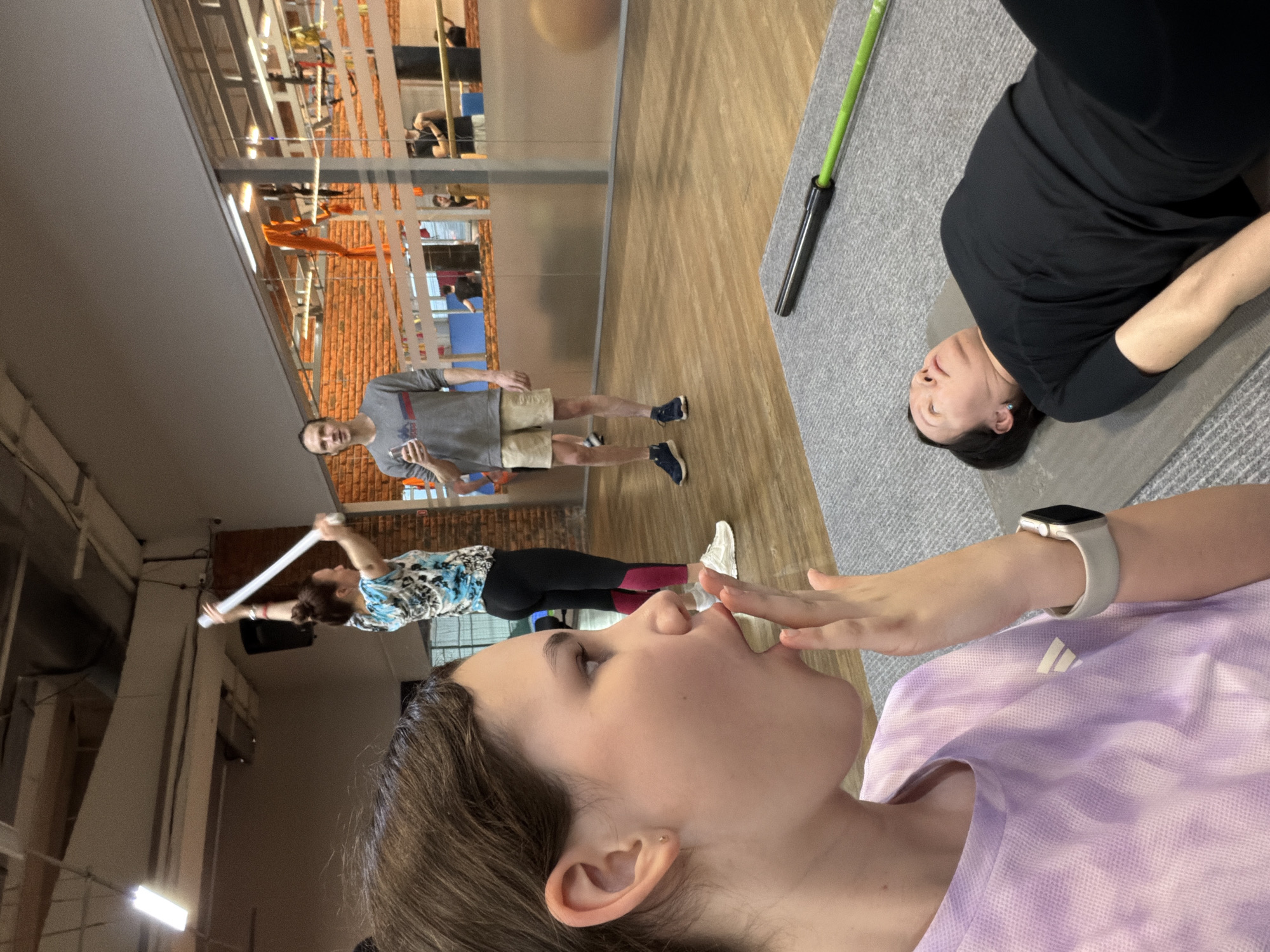 A selfie-style image is captured indoors, potentially at a fitness center in Chaikovsky, Russia. The foreground features a young woman in a light purple Adidas t-shirt, kissing her hand. In the midground, other people are present, including a man holding a white pole, a woman in a printed top, and a man in a grey t-shirt and beige shorts. A woman in black attire lies on a grey mat in the background. The environment includes fitness equipment, wooden floors, and a mirrored wall that reflects the scene, possibly revealing the gym's interior.