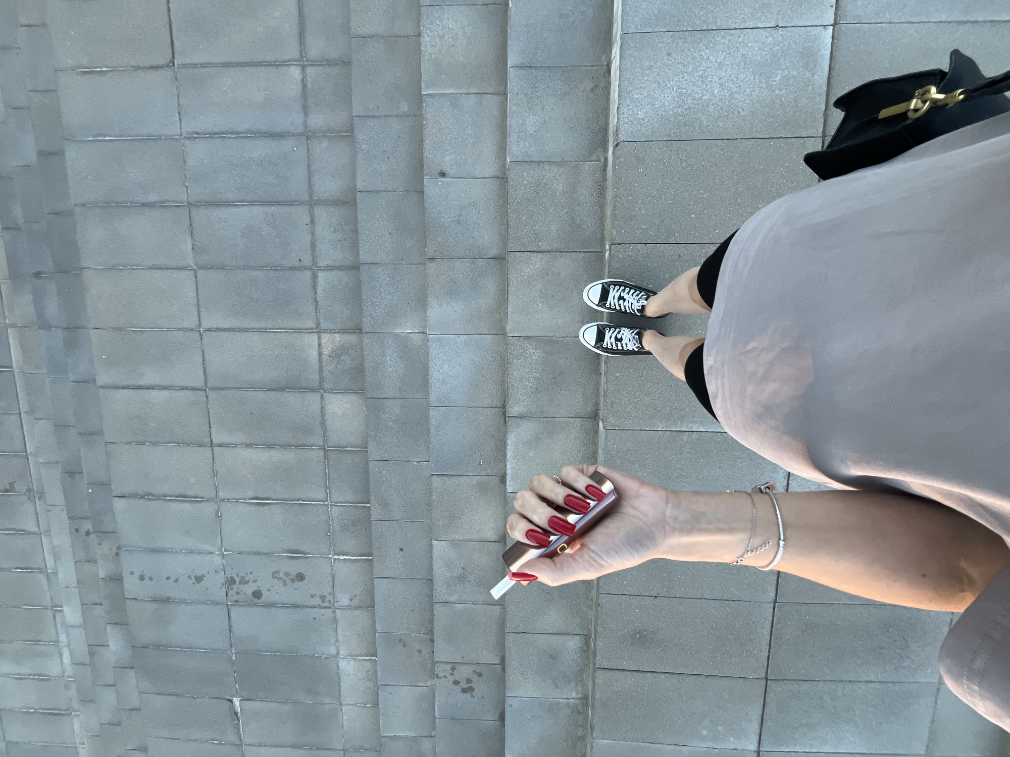 The media file shows a first-person perspective of a person standing on a grey, tiled staircase outdoors. The person is wearing black sneakers with white laces and black shorts, along with a light-colored, flowing dress or skirt. A black handbag with gold hardware is visible near the person's hip. In the foreground, the person's hand, adorned with long, red-painted fingernails and a silver bracelet, is holding a metallic electronic cigarette device. The overall scene suggests a casual setting, possibly a public space or walkway with stairs. The lighting indicates it could be daytime, with no specific weather conditions apparent. The photo is taken from a high angle, looking down at the person's feet and hand. There is no visible text in the image. The location is specified as Nizhny Novgorod, Russia, but this context is not directly evident from the visual elements alone.