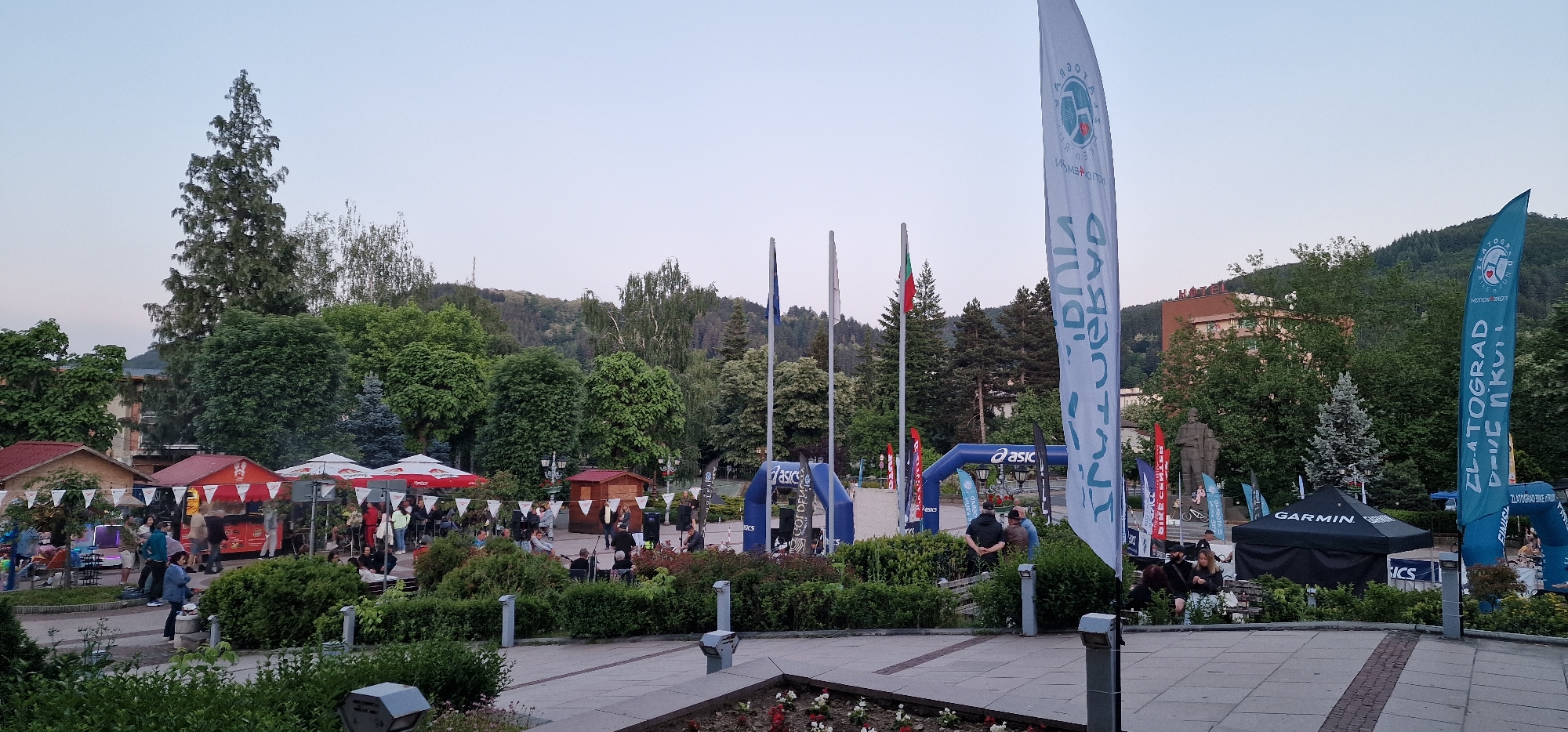 The image shows a public space in Zlatograd, Bulgaria, likely a town square. There are food stalls with red and white umbrellas on the left, suggesting a market or event. Several people are present, mostly standing and walking, implying social interaction. There are flags bearing local symbols and advertisements for brands like Asics and Garmin. An archway also bears the Asics logo, suggesting a sporting event. The Bulgarian national flag is visible. There's a statue in the background on the right. Green trees fill the background. The sky is light, suggesting dusk or dawn.