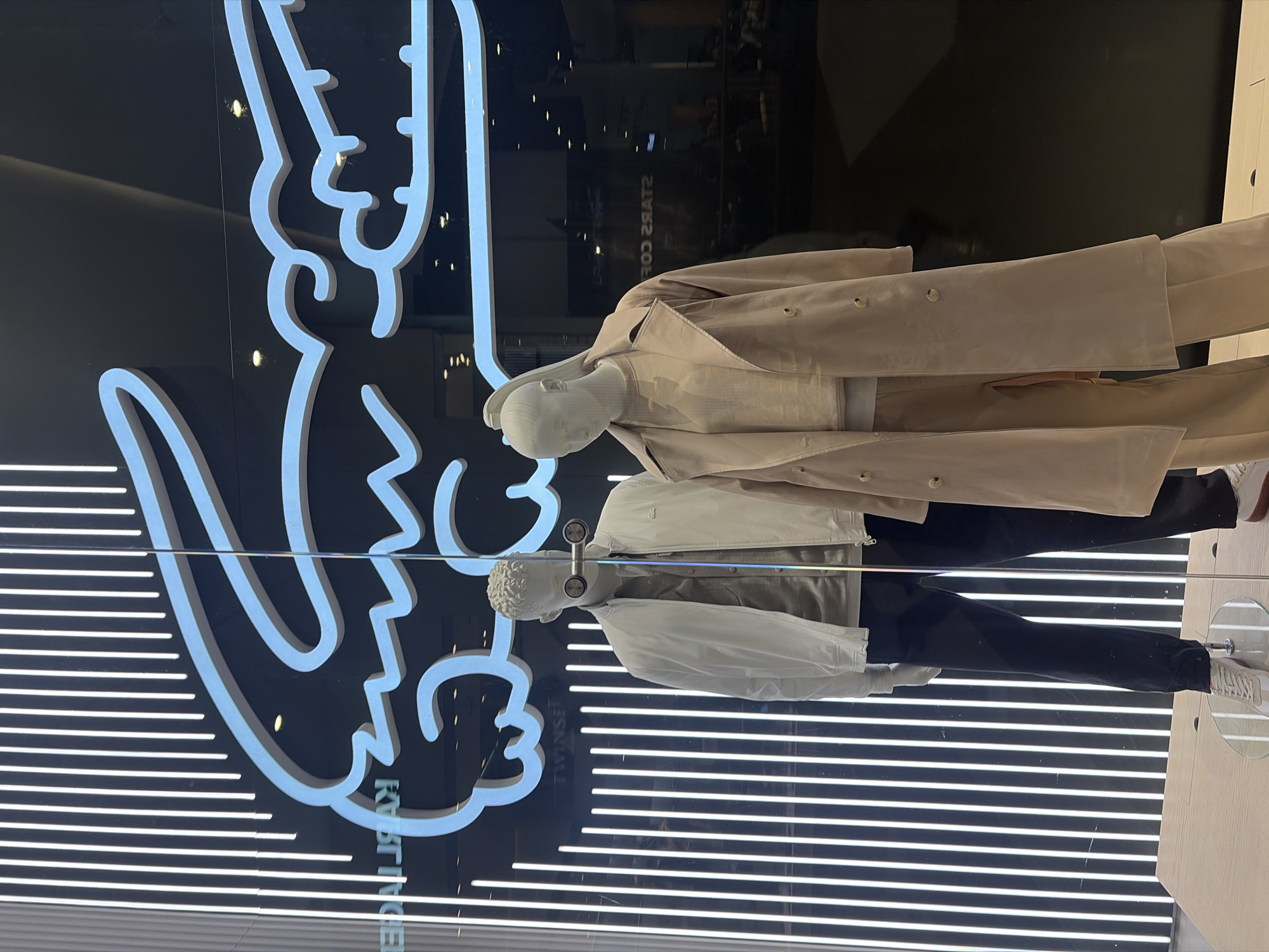 Here's a detailed description of the image:
**Overall Impression:** The image captures a storefront display, likely for a clothing brand, featuring mannequins and a large logo.
**Specific Details:**
* **Location:** The text "KVARTIRINGEI" is visible. This indicates that the photo was taken at the Galeria shopping center in Saint Petersburg, Russia.
* **Subjects:** Two mannequins, both with textured heads, are dressed in contemporary clothing.
* The top mannequin wears a light tan trench coat over a lighter-colored top and trousers.
* The bottom mannequin wears a white jacket over a lighter-colored top and dark trousers. White sneakers are visible on this mannequin.
* **Objects:**
* A large, white neon logo with a stylized, elongated design is prominently displayed behind the mannequins.
* Horizontal white lines are visible in the background, possibly part of the display or the building's design.
* The floor surface appears to be a light color, possibly wood or laminate.
* **Environment:** The display is situated behind a large glass window, creating reflections. The light in the scene appears to be diffused, suggesting indoor lighting.
* **Actions/Interactions:** The mannequins are posed statically, showcasing the clothing.
**Key Elements:** Clothing display, mannequins, Saint Petersburg, clothing store, Galeria shopping center.