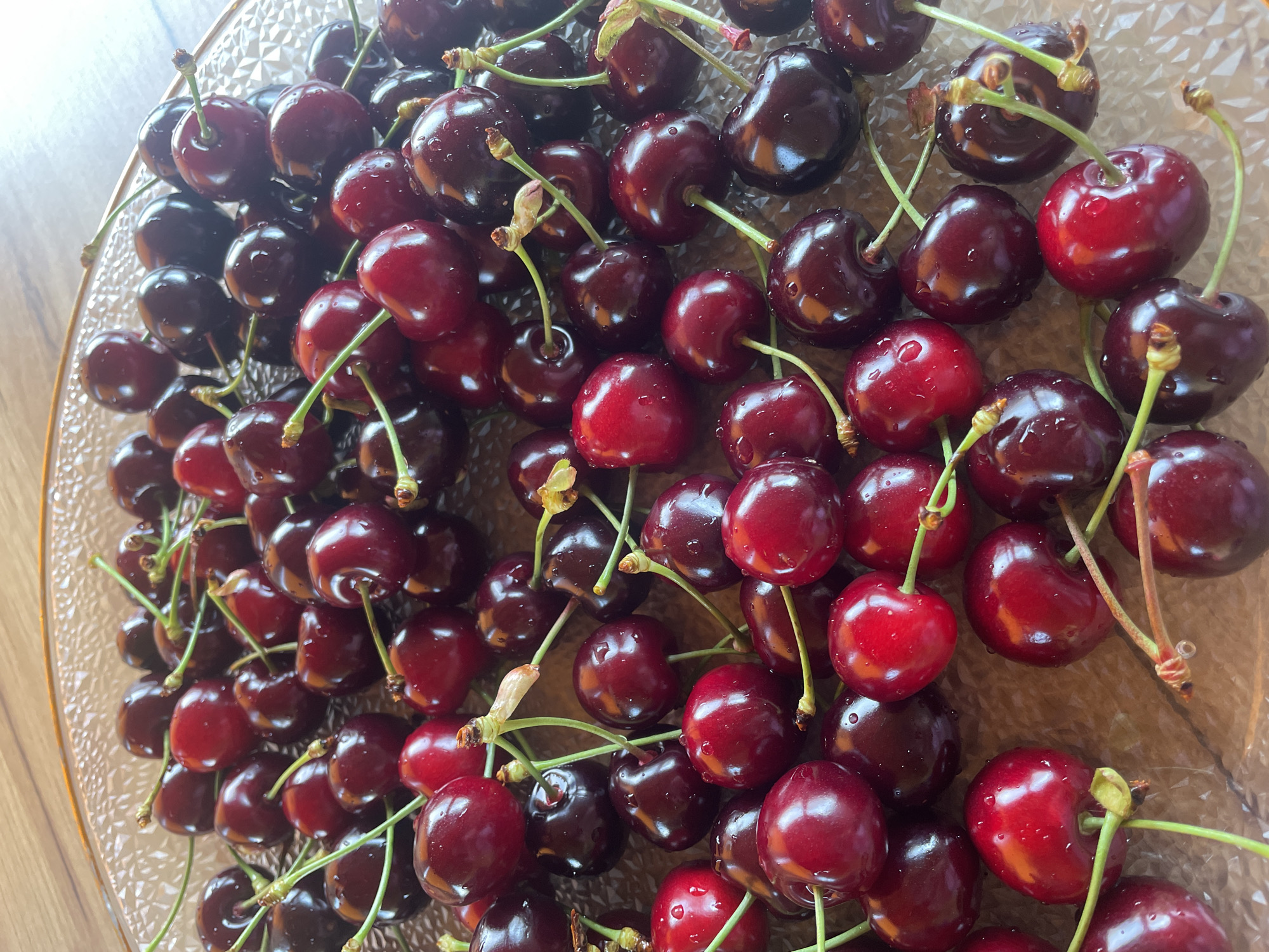A high-angle, close-up photograph displays a collection of approximately 50-70 fresh cherries arranged within a clear, textured glass plate. The cherries exhibit varying shades of red, ranging from a vibrant ruby red to a deeper, almost black-red hue. Numerous small water droplets are visible on the skin of many cherries, suggesting recent washing or freshness. Most cherries retain their green stems, often clustered, with some showing light brown calyx remnants at their attachment points. The glass plate features a raised, faceted, or dimpled pattern on its surface, causing light refraction. The rim of the plate, visible at the top and right edges, appears to be made of glass with a slight amber or golden tint due to reflections. The plate rests on a smooth, polished wooden surface, visible as a warm brown background with subtle grain patterns. The scene is evenly illuminated, producing highlights on the wet, glossy cherry skins. There are no people, explicit actions, or interactions depicted.