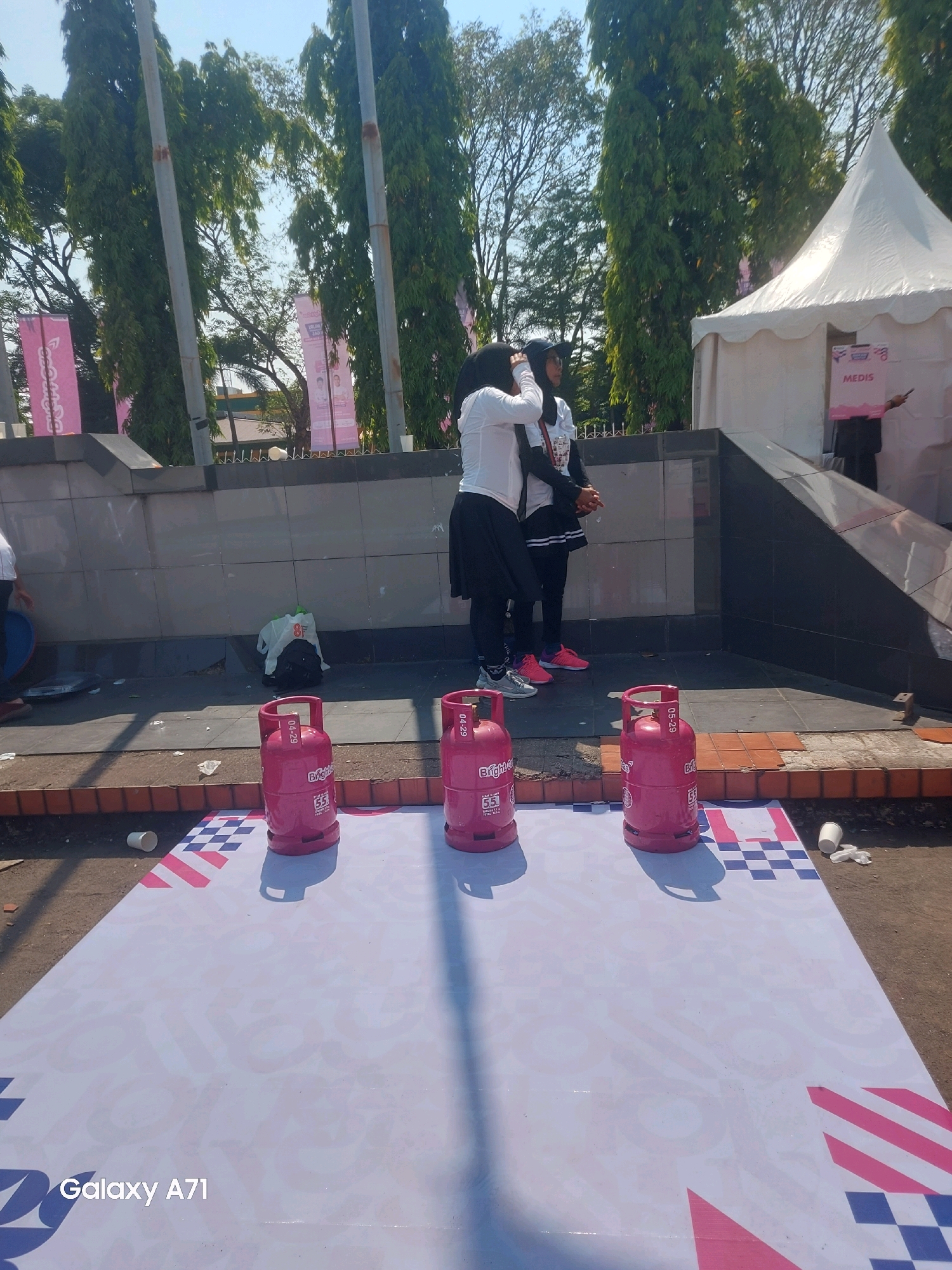 This is a medium shot, outdoor, eye-level, full-color photograph taken on a sunny day. The scene appears to be an outdoor event or promotional activity. In the foreground, there are three pink LPG gas cylinders placed on a large white banner with a checkered pattern in blue and pink. The banner has some faint text and logos on it. One of the cylinders has "Bright Gas" and "5.5" visible on its side. Two white cups and some crumpled paper are scattered on the ground near the banner.
Behind the banner, a stone wall separates the foreground from the middle ground. A black backpack and a white plastic bag are placed near the wall. In the middle ground, two women are standing, both wearing dark hijabs and white tops. One woman is wearing a black skirt and athletic shoes, while the other is wearing athletic shorts and sneakers. They appear to be engaged in a conversation or interacting with each other.
In the background, there are tall trees with green foliage, suggesting a park-like setting or an area with significant greenery. Behind the trees, a white tent is visible, possibly a booth or stage for the event. A sign on the tent reads "MEDIS" in pink lettering. A person in a dark outfit is partially visible standing in front of the tent, holding a pink sign and pointing. Pink banners with text are also visible hanging between poles in front of the trees. The overall environment is bright and appears to be a public outdoor space. The presence of promotional materials, tents, and people dressed casually suggests a public event or gathering. The branding on the LPG cylinders indicates they might be related to a fuel company or a promotion involving gas services.