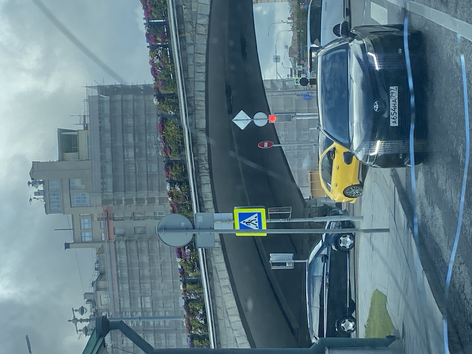 In an urban setting under an overcast sky, a black Toyota Camry, license plate A654HA 797, is stopped at a pedestrian crossing. The vehicle's headlights are on. To the left of the Camry, a bright yellow hatchback is also present. Behind these vehicles, a dark-colored van is visible. Further in the background, an arched stone bridge spans over the roadway, decorated with planters filled with pink and purple flowers. A building undergoing construction, covered in scaffolding, stands behind the bridge. Traffic lights and pedestrian crossing signs are also visible. The road surface appears wet.