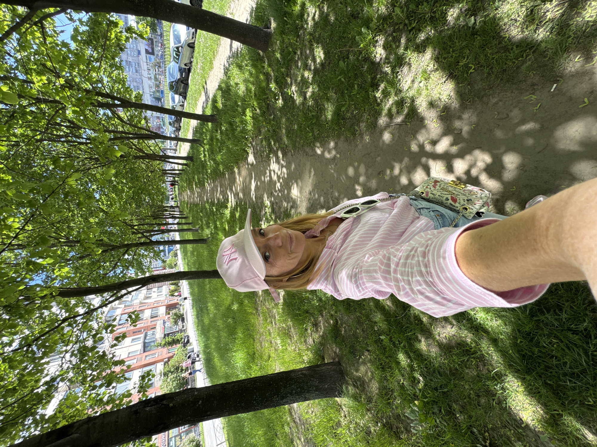 The image shows a woman taking a selfie outdoors on a bright, sunny day. The woman is positioned in the foreground, looking directly at the camera with a relaxed, slightly smiling expression. She has blonde or light brown hair and is wearing a pink and white striped long-sleeve shirt, light-colored jeans, and a pink baseball cap with a prominent white "NY" logo. Sunglasses are perched on top of her head, and a small, light-colored handbag with a patterned design (featuring elements like lips and crowns) and a gold chain strap is visible by her side.
The scene depicts a vibrant green park or urban greenway, characterized by a long row of mature trees with lush foliage, casting dappled shadows across a dirt path and grassy verges. The bright sunlight indicates a clear mid-day or afternoon. In the background, beyond the trees, multi-story residential or commercial buildings rise, typical of an urban landscape. A line of parked cars is visible along a street bordering the park. Faint figures of other people can be seen further along the path. The setting suggests an urban park in Saint Petersburg, Russia.
The only clearly legible text is the "NY" logo on the baseball cap. Some very blurry Cyrillic text can be faintly discerned on distant buildings, appearing to be commercial signs, but is largely illegible due to distance and resolution.