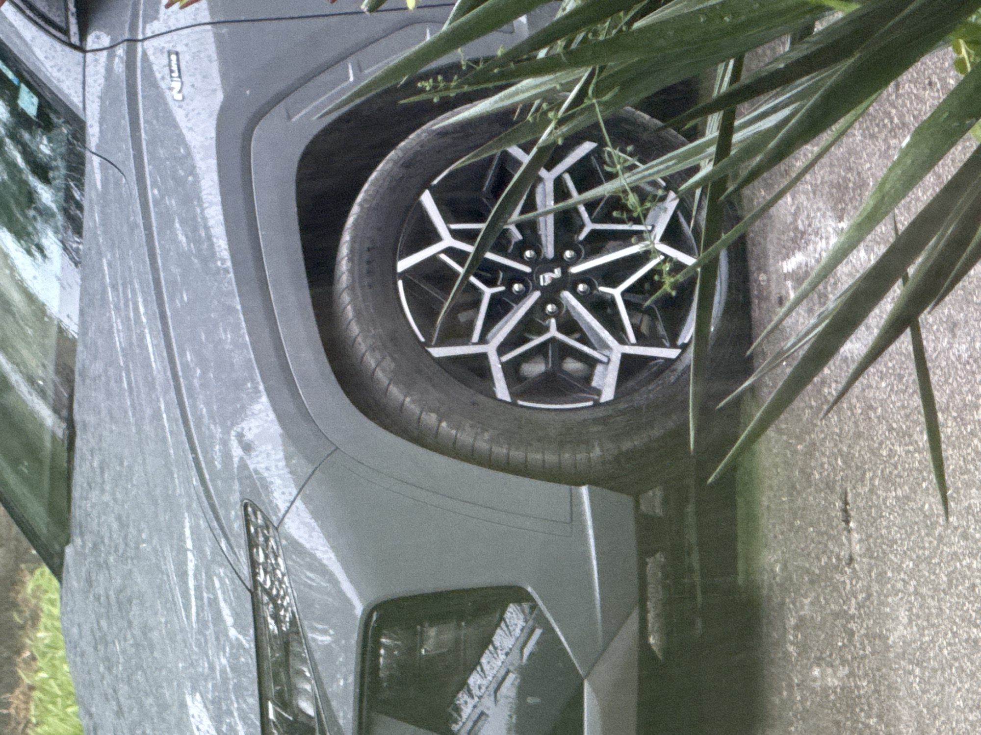 A gray Hyundai Kona N is parked on a concrete surface. A tire and wheel are visible. Green foliage is partially obscuring the tire. The "N" logo is visible on the wheel's center cap. The backdrop reflects buildings. No people are visible. Location is indeterminate.