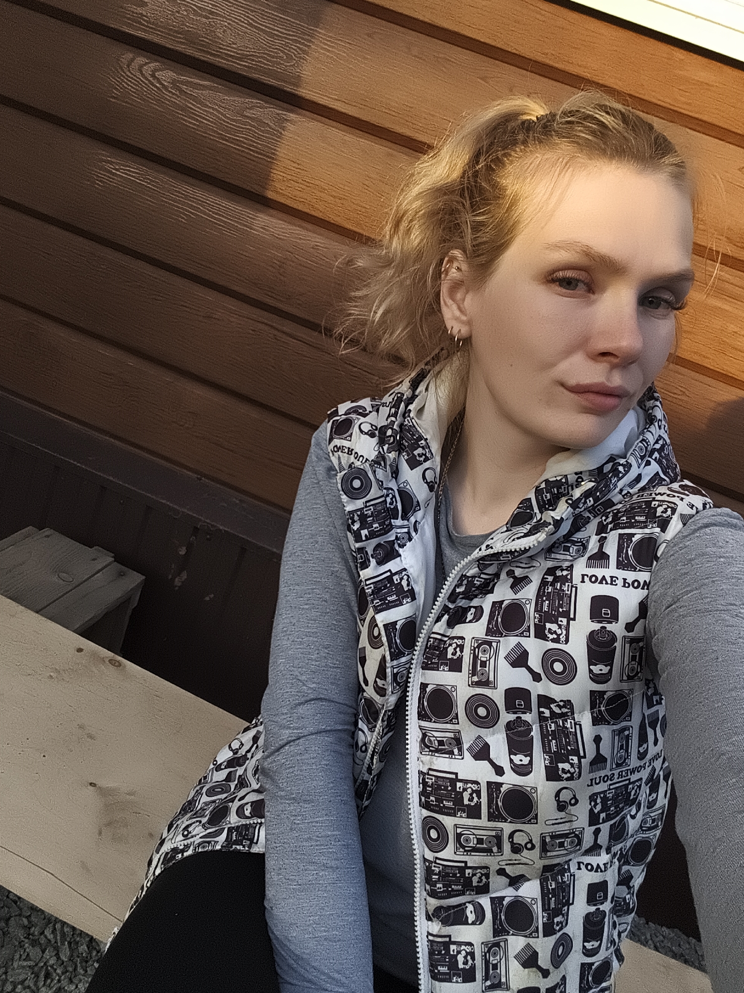 A blonde woman is sitting on a wooden surface in Русский Вожой, Russia. She is wearing a grey long-sleeved shirt and a white vest with black drawings. The background is a wooden wall with horizontal planks.