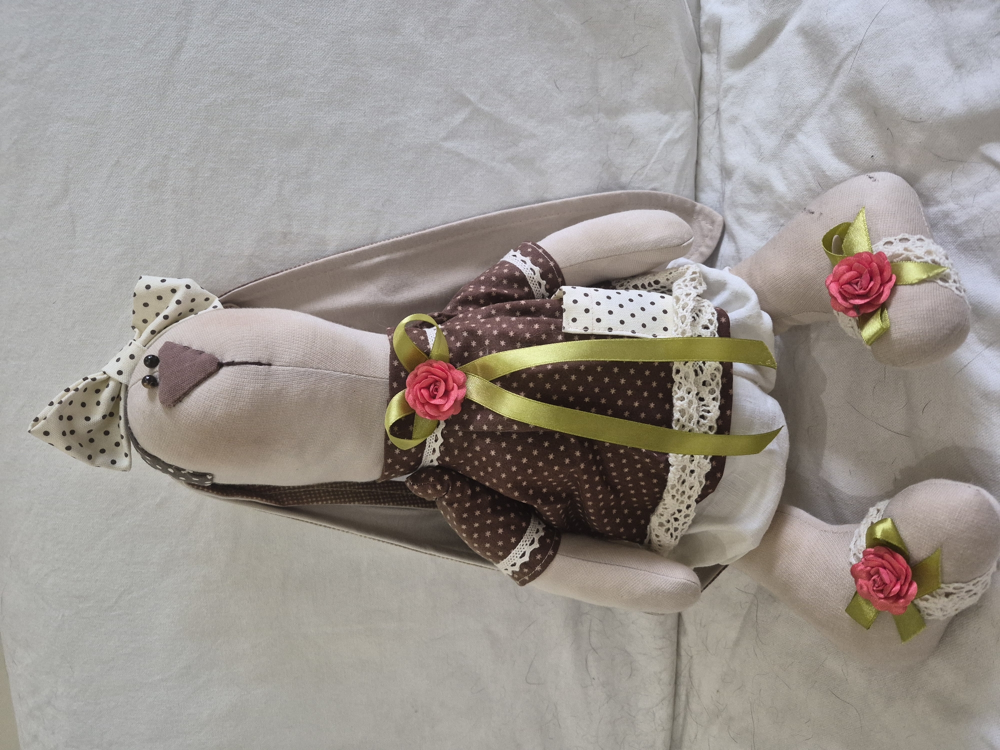A stuffed bunny toy is sitting upright on a light-colored, textured surface, possibly a sofa or bed. The bunny is made of light brown fabric and has long, floppy ears lined with brown fabric. It wears a white ruffled bloomers, a brown polka-dotted shirt with white lace trim at the cuffs and hem, and a cream-colored bow with dark brown polka dots on its head. A lime green ribbon with a pink fabric rose is tied around the bunny's neck, with the ribbon hanging down the front of its shirt. Each foot of the bunny is decorated with a similar lime green ribbon and pink fabric rose, also accented with white lace. The bunny's eyes are black buttons, and its nose is a small brown fabric triangle. The background is a neutral, textured fabric. There is no indication of a specific city or location within Vlast Truda, Russia, or any human interaction.