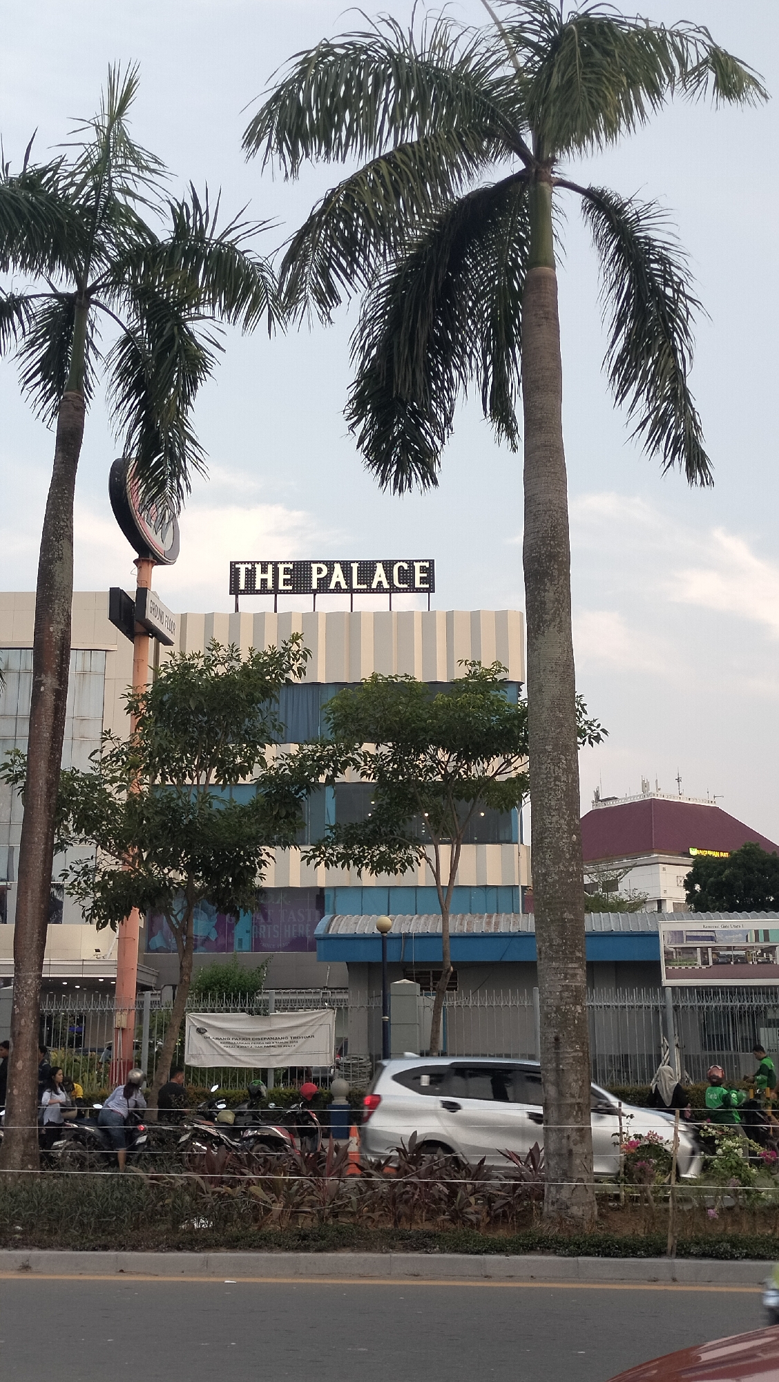 This image depicts a street scene in Batam, Indonesia, likely in the vicinity of Nagoya Hill Shopping Mall, given the signage. A tall palm tree is prominent in the foreground, obscuring part of a building. The building features the illuminated sign "THE PALACE" at the top. Below the sign, there are multiple levels of windows and a light blue facade. In front of the building, there is a metal fence and a row of parked motorcycles, with several people standing around them. A silver car is driving past on the road, which is in the lower portion of the image. Other smaller trees and plants are visible along the roadside and in front of the building. In the background, there are more buildings, including one with a distinctive red roof. The sky is overcast.