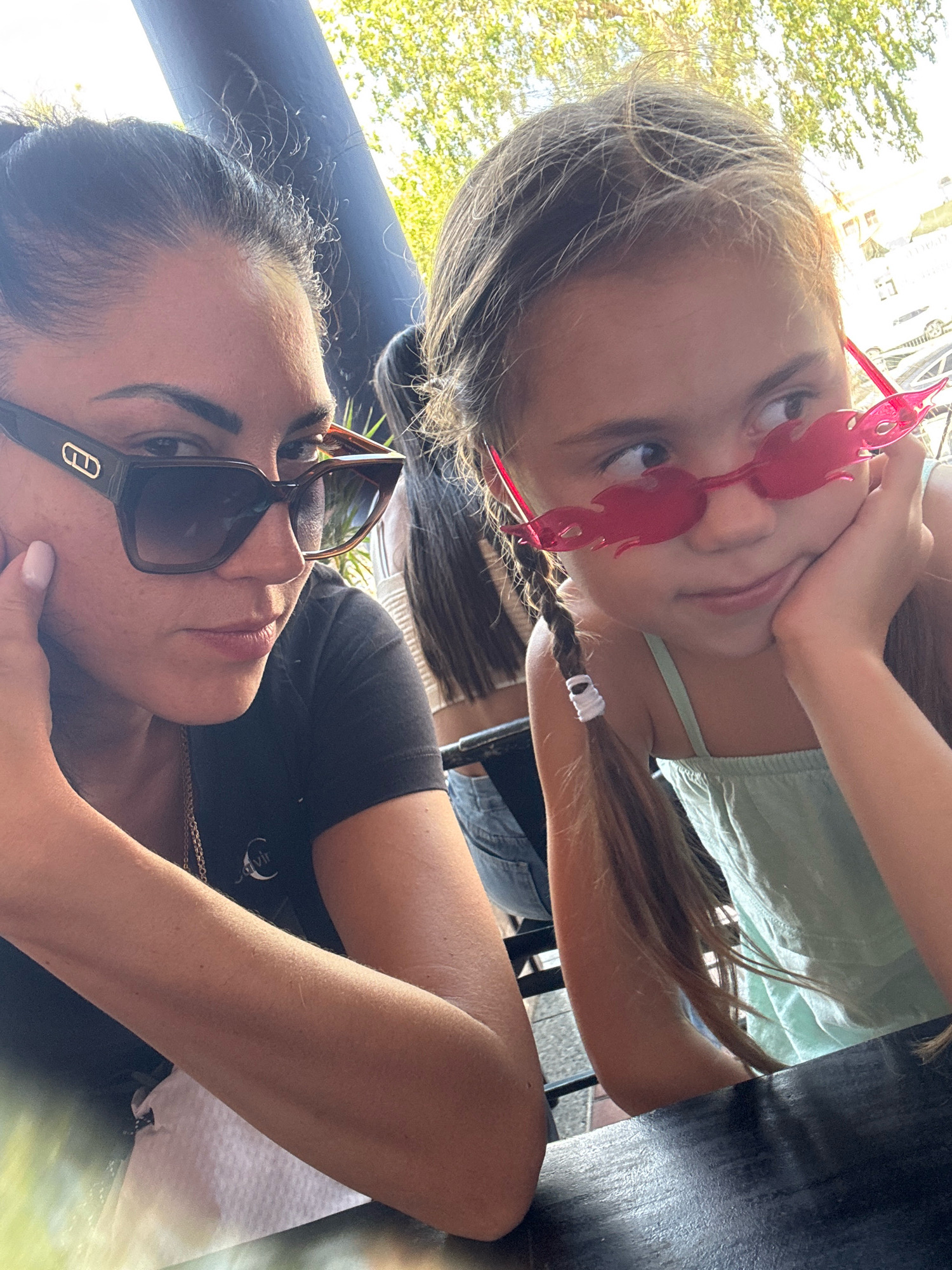 This is a medium shot photograph taken outdoors in Netanya, Israel, featuring two females at a table. The female on the left, an adult with dark hair pulled back, is wearing dark-rimmed sunglasses and a black t-shirt. Her left arm is bent, and her left hand rests on her cheek. To her right is a young female with long, braided blonde hair, also wearing sunglasses, these ones red and flame-shaped. She is wearing a light-colored sleeveless top and has her right hand on her chin, resting her elbow on the table. The background shows a dark pillar, trees with green foliage, and a building with windows, suggesting an urban or semi-urban outdoor setting. The lighting indicates it is daytime.