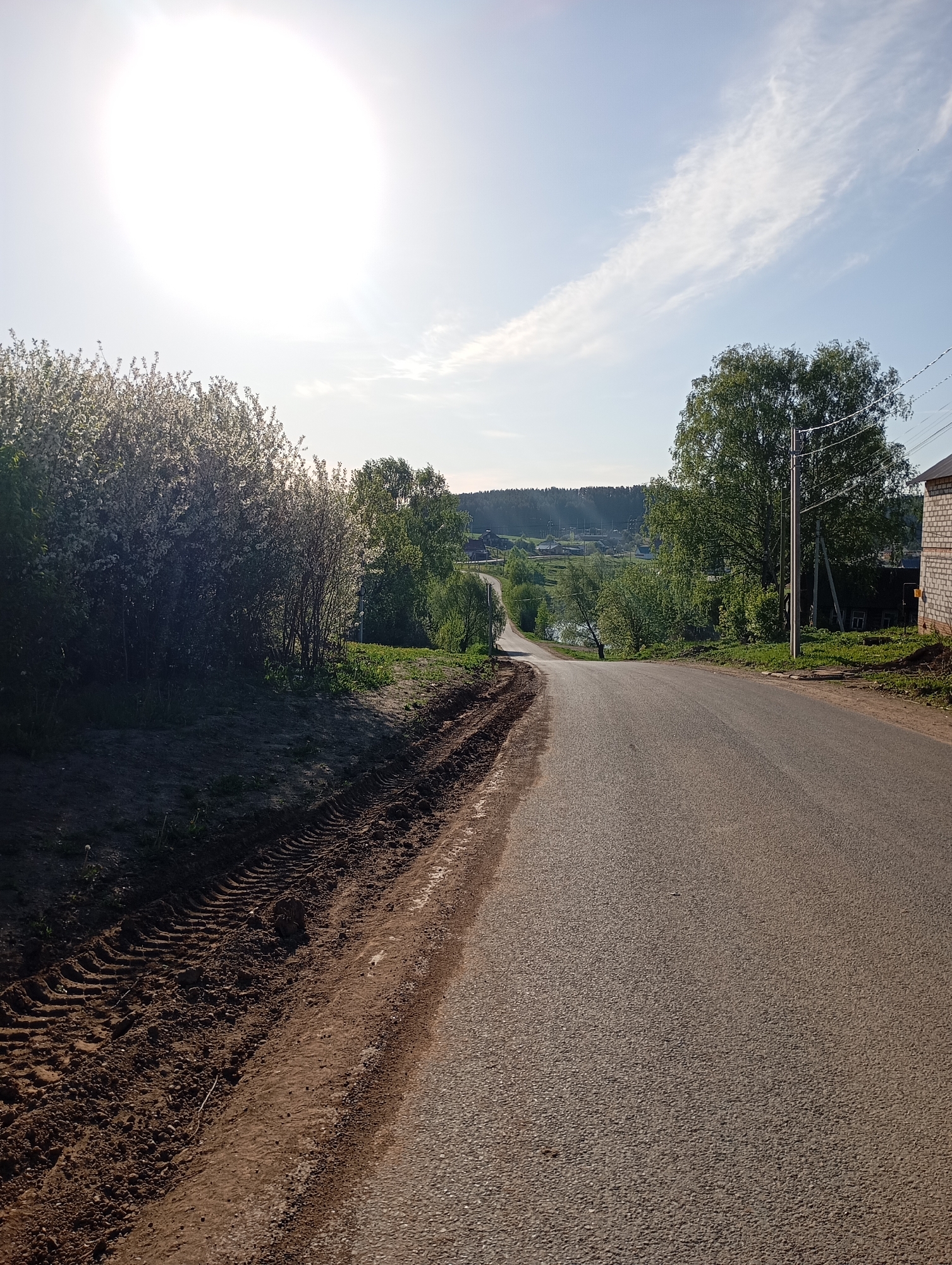 A paved road winds through a rural landscape on a sunny day. The road slopes downward, leading toward a distant view of a body of water surrounded by trees and houses. On the left side of the road, there is a stretch of dirt with clear tire tracks. A lush bush with small white flowers grows along the edge of the road. On the right side, a utility pole stands near a building. The sky is bright with a large sun and some wispy clouds. The overall scene suggests a quiet, countryside setting. The location is in the city of Unknown, Unknown.
