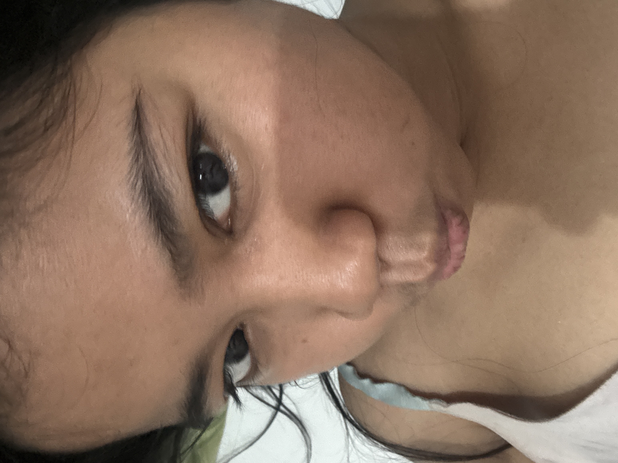 The image is a close-up, self-taken portrait of a young person's face, with the camera positioned slightly above and to the side. The subject has dark hair and dark eyes, and is making a puckered expression with their lips. Their skin tone is medium brown. They are wearing a light-colored tank top with thin straps. The background is out of focus and appears to be a plain, light-colored wall or surface, with a hint of green to the left. There are no visible elements that would indicate a specific location in North Jakarta, Indonesia. The lighting appears to be indoor, with a slight shadow across the bridge of the nose and cheek.