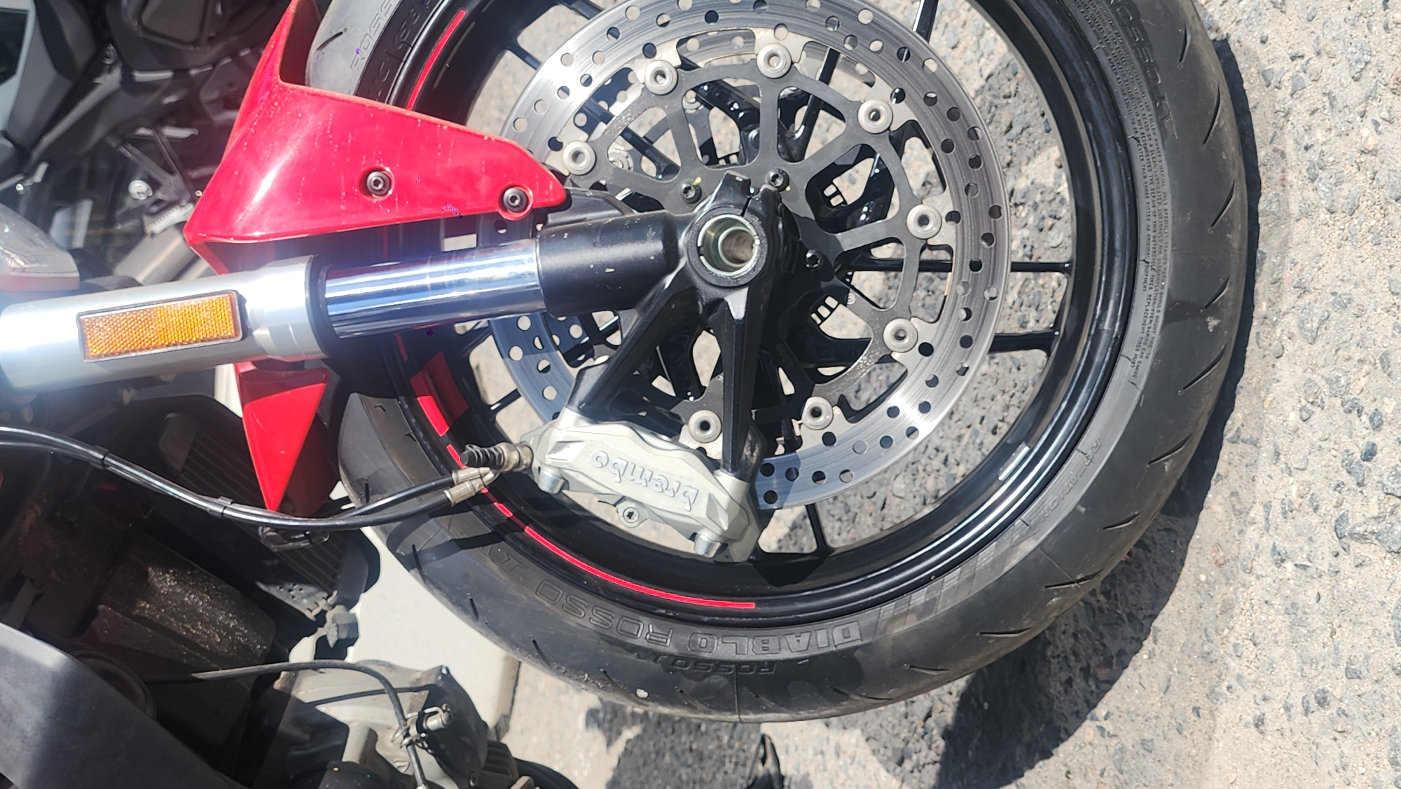 The image shows a close-up of the front wheel of a red motorcycle parked outdoors on a paved surface that appears to be asphalt or concrete, with small gravel present. The motorcycle has a black wheel rim with a red stripe, a silver Brembo brake caliper, and a perforated brake disc. The tire is a Pirelli Diablo Rosso. Part of the red fairing, the front suspension fork, and a reflective orange marker are visible. The background is blurred but suggests other vehicles or structures. There are no people visible in the image. There is no information or indication to suggest this location is in Moscow, Russia.