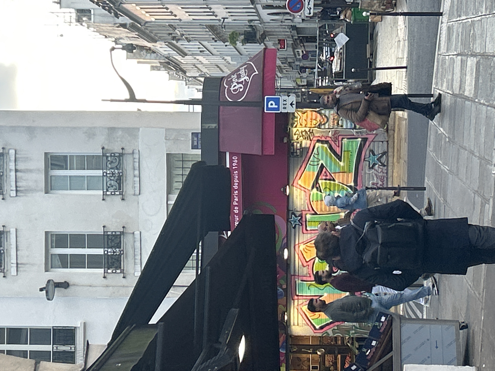 The image shows a street scene in Paris, France. In the foreground, there are several people walking along the sidewalk. There is a shop front with graffiti on it. A red awning displays a logo and the text "de Paris depuis 1960." A parking sign with an arrow pointing towards "REX" is visible. Across the street, there is a building with windows. A street lamp is also visible, as well as traffic lights and cars further down the street. The sky is overcast.