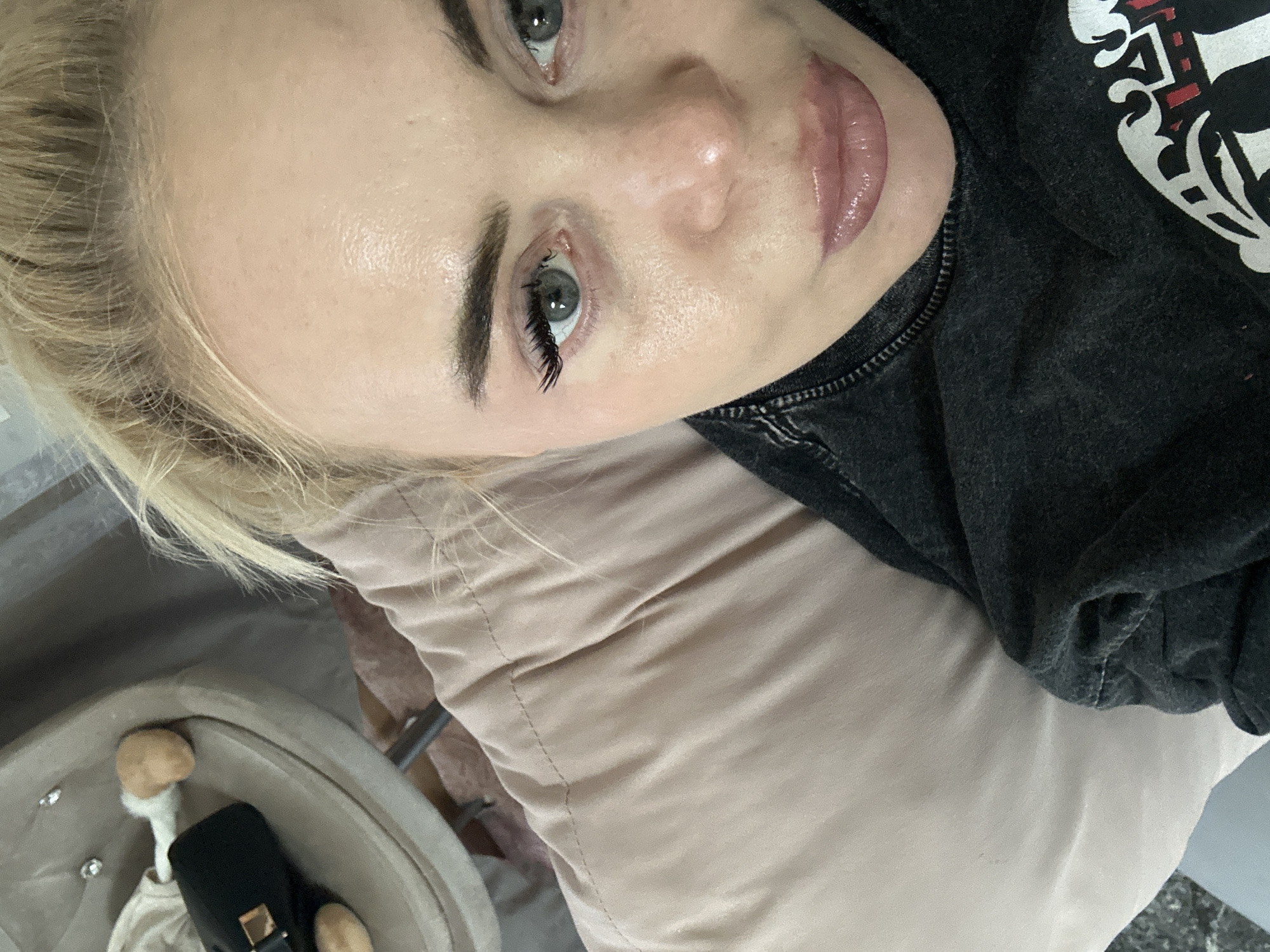 A fair-skinned woman with blonde hair, light gray eyes, and visible makeup (eyelash extensions, lip gloss) is looking upward at the camera. She's wearing a dark-colored shirt with a white design. She appears to be lying down on a light-colored pillow or cushion. A chair with a bag on it is visible in the lower-left corner of the image. The location is indicated as Высокий, Russia, but there are no visual elements in the image to confirm this location.