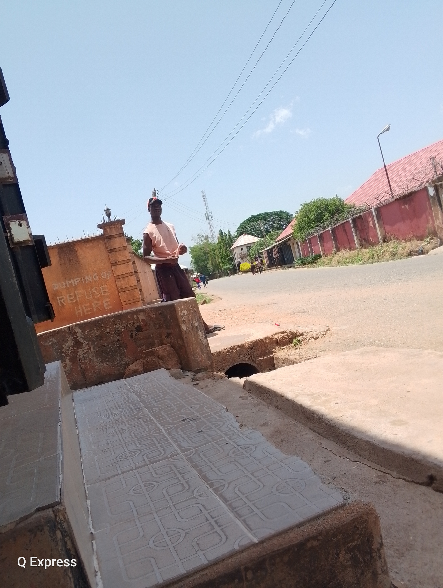 A person is standing on a stone structure on a roadside in Gura Top, Nigeria. The structure is marked with the words "DUMPING OF REFUSE HERE." The surrounding environment includes a street, buildings with red roofs, a fence topped with barbed wire, and utility poles. The weather appears sunny.