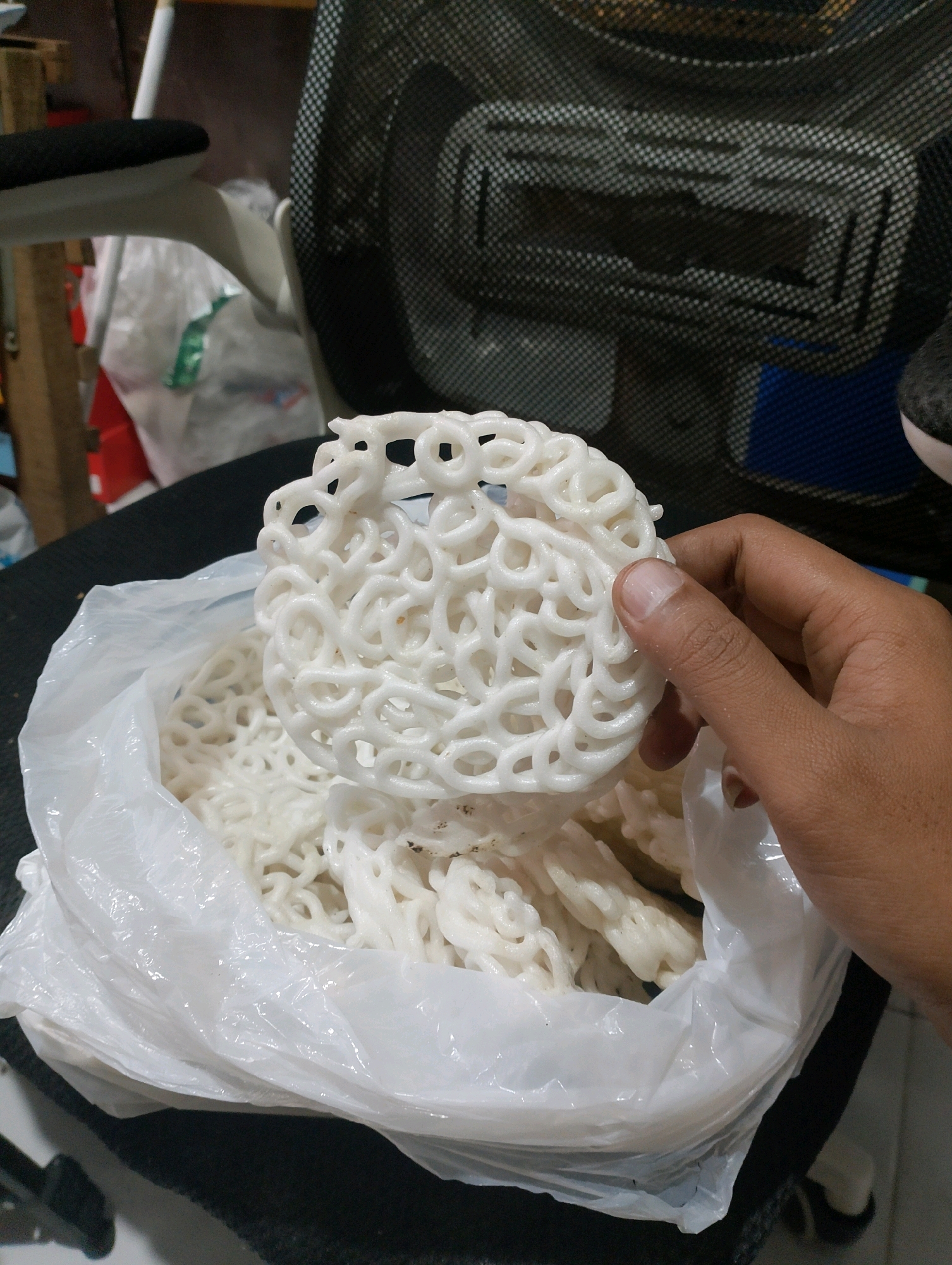 A person's hand is holding a white, circular cracker made of intertwined loops, above a plastic bag filled with more of the same crackers. The crackers are a light white color and appear to be uncooked. The bag is translucent and somewhat crumpled. In the background, a black mesh chair is visible, along with some wooden objects and plastic sheeting. The scene appears to be indoors, possibly in a home or storage area, with the context suggesting the location is Sumberjati, Indonesia.