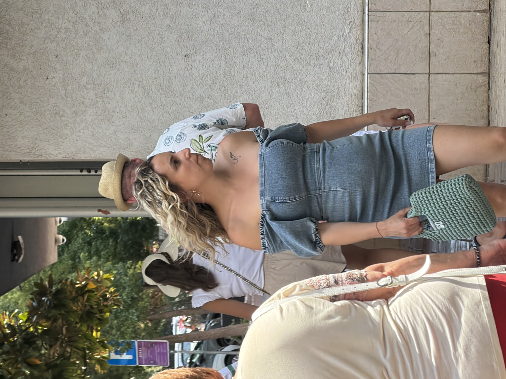 The image depicts a street scene in Budva, Montenegro. A woman with blonde, curly hair is walking forward, wearing an off-the-shoulder denim dress and carrying a green crocheted handbag. She has a tattoo on her left shoulder blade. Behind her, a man wearing a straw hat and a patterned shirt is partially visible. To the left, another woman is seen from behind, wearing a light-colored top and carrying a white bag. In the background, trees and a blue parking sign are visible. The ground is paved with tiles. The overall lighting suggests it is daytime, possibly late morning or early afternoon.