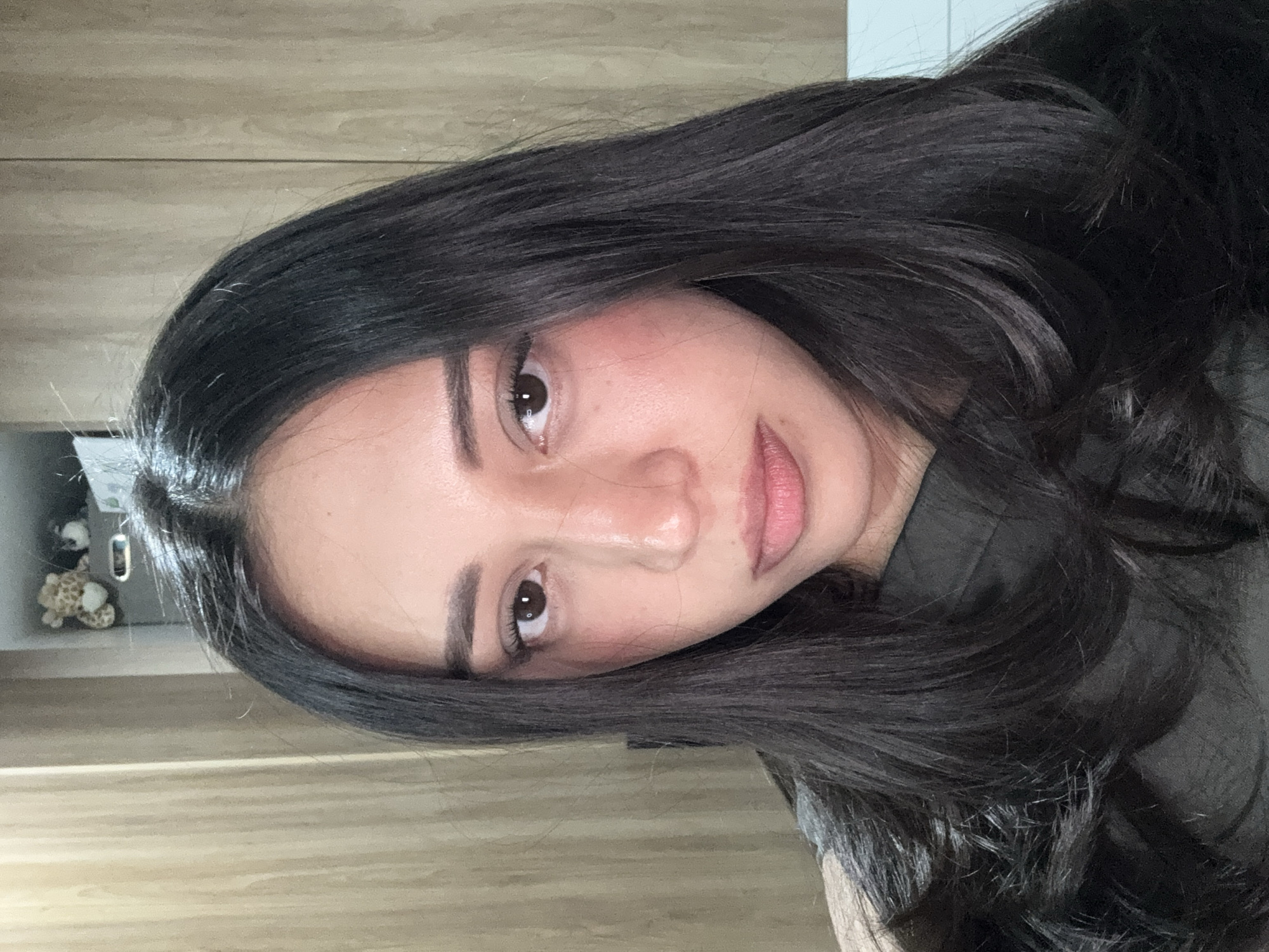 A frontal portrait shot captures a female subject, approximately 20-30 years old, looking directly at the camera. She possesses dark, long, wavy hair, parted centrally, extending over her shoulders. Her eyes are dark brown, and her eyebrows are well-defined. Her skin tone is light to medium, exhibiting a slight reddish tint on the cheeks. Her mouth is slightly agape, maintaining a neutral expression. She is wearing a dark, possibly black or dark gray, garment with a visible neckline. The environment is an indoor setting. The background features light-toned wooden paneling or furniture on both the left and right sides, suggesting a wall or cabinet. In the upper right background, a shelf is visible, containing a small, light-colored stuffed animal with brown spots (resembling a giraffe) and a smaller, darker stuffed animal or object next to it, along with a gray storage container. The location within the city of Puebla, Mexico, cannot be determined from the visual information present in the image. No discernible actions or interactions beyond posing for the photograph are evident.