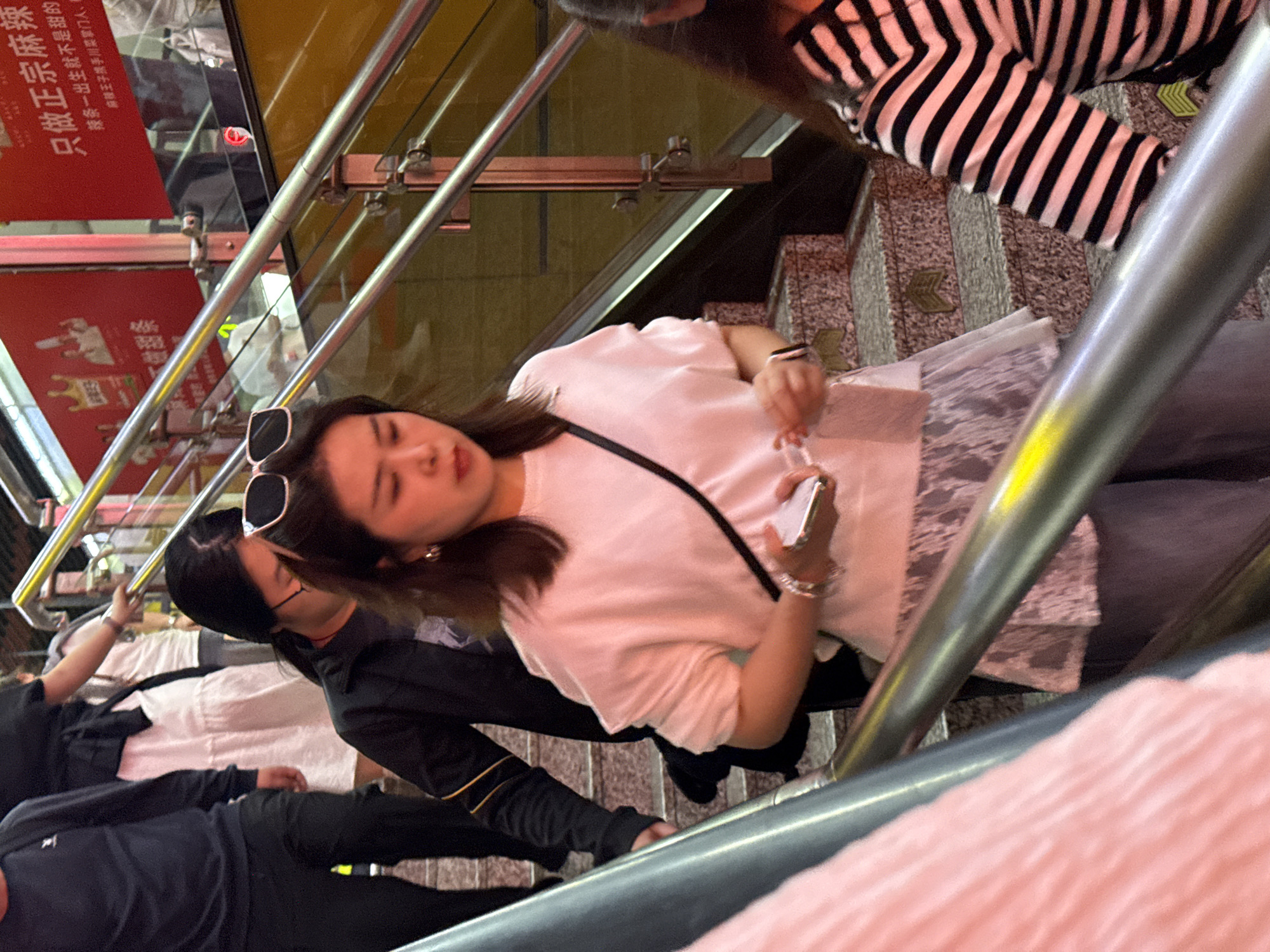 Observed at a canted angle, the image shows individuals on an escalator within what appears to be a commercial area of Yuzhong District, China. The backdrop features advertising signage in Chinese.
In the foreground, individuals are ascending on the escalator. A woman in a white top, sunglasses perched on her head, is holding a mobile phone. Another person is situated to her left, wearing a white shirt and a dark jacket. A third person is visible at the bottom left of the frame, clad in a dark blue or black top and pants.
The escalator features a metallic handrail. A person dressed in a black and white striped top is also visible. The scene appears to be captured at night. The lighting is predominantly artificial.