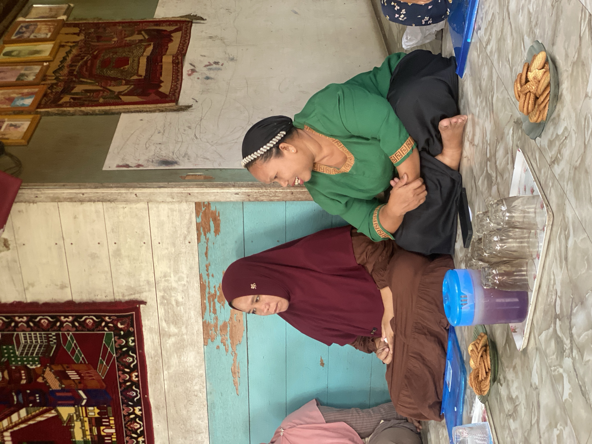 Two women are sitting on the floor indoors, in what appears to be a room in Bawahan Selan, Indonesia. The woman on the left is wearing a maroon hijab and a brown dress, and is looking to the left. The woman on the right is wearing a black headscarf with a beaded headband and a green top with gold trim, and is looking to the right and smiling. They are both sitting with their legs crossed in front of them. In the foreground, on a tray, are several glasses, a blue pitcher filled with a purple liquid, and a plate of cookies. In the background, the wall is made of light-colored wood planks painted turquoise on the lower half. There are two woven wall hangings visible, one on the left wall and one on the right wall, along with several framed pictures above the right wall hanging. The floor is tiled with a marble-like pattern.