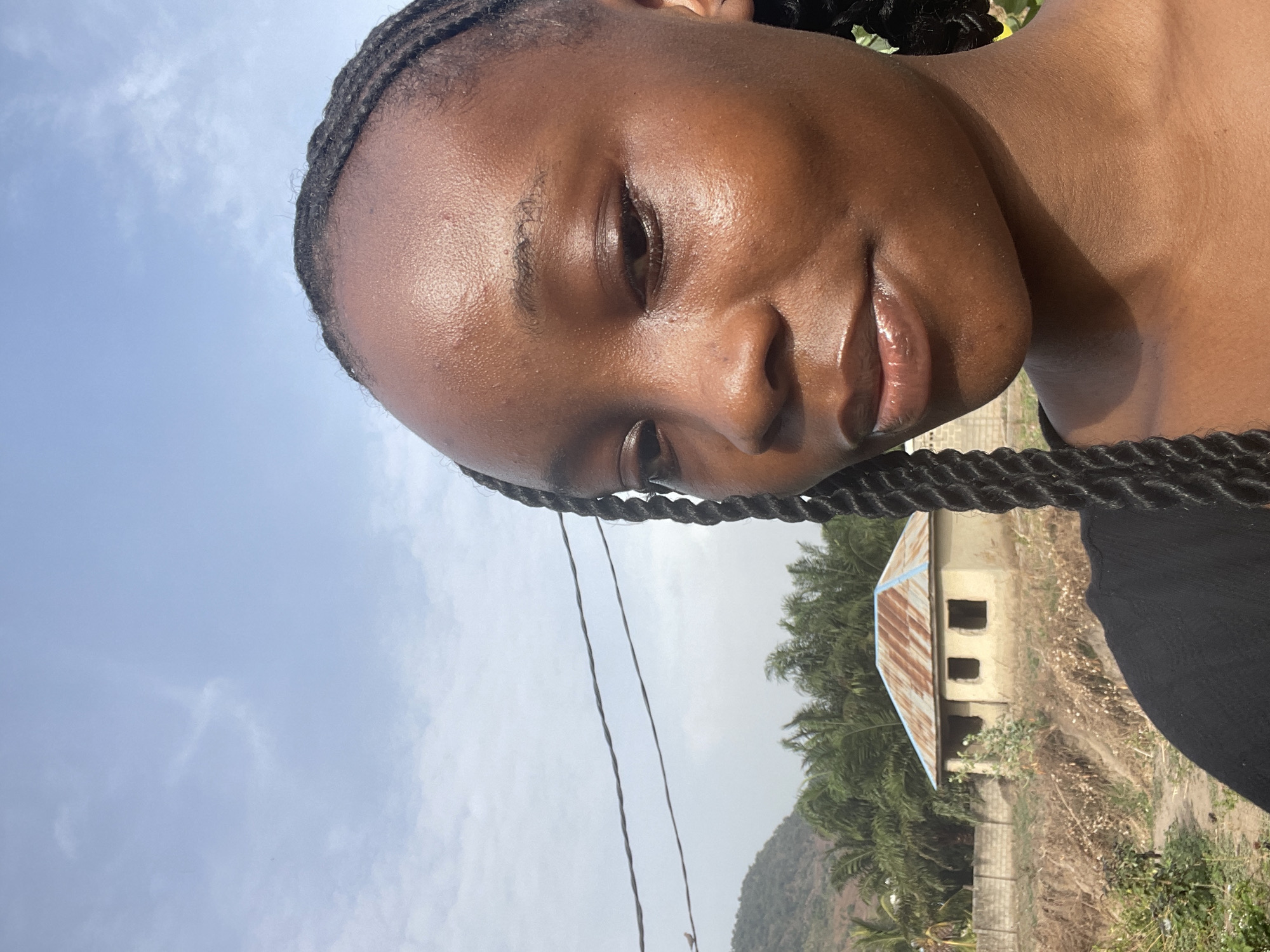 A rotated selfie captures a young person with dark skin and braided hair, smiling gently towards the viewer. The person's face, radiant under bright sunlight, fills much of the frame. Their lips are moisturized, and their dark eyes gaze slightly past the camera. Braids are visible along their hairline and resting on their shoulder.
The background reveals a clear, light blue sky with a few faint clouds, indicating a bright daytime setting, likely in the morning or afternoon. Below the horizon line, a landscape typical of Lokoja, Nigeria, unfolds. Lush green palm trees stand alongside dry, brownish vegetation, with rolling hills or mountains visible in the distance.
In the middle ground, a simple, unfinished building features a weathered, rusty corrugated metal roof and empty window openings. A block wall or fence is discernible nearby. Two dark wires or cables stretch vertically through the left side of the frame. The overall impression is one of a serene, sunny day in a natural, semi-rural environment. No text is visible in the image.