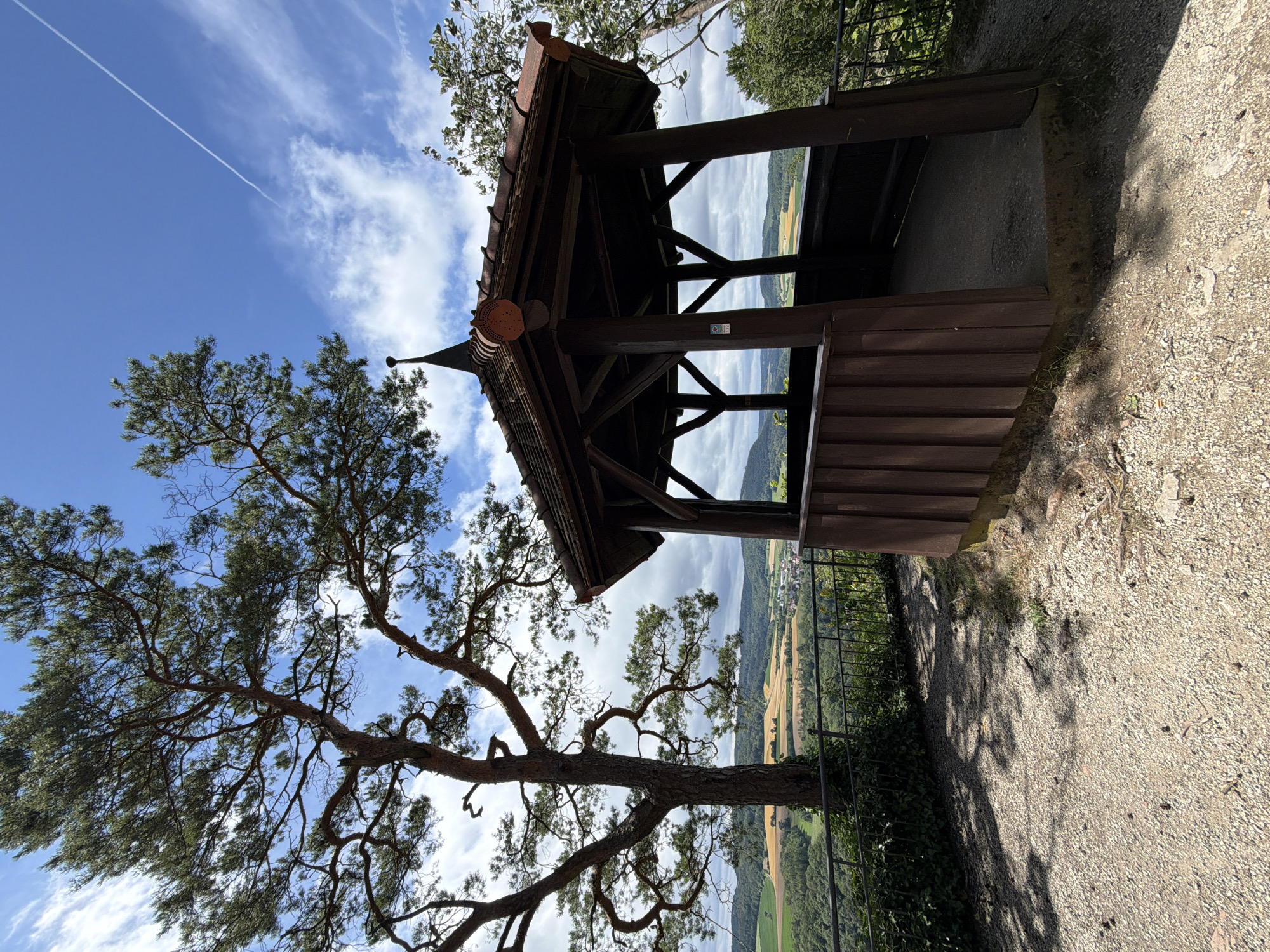 A wooden gazebo with a dark brown, tiled roof sits on a gravelly overlook, offering a view of a rolling, green landscape under a blue sky with scattered clouds and a distant contrail. The gazebo is octagonal, with vertical wooden planks forming its lower walls and open upper sections supported by wooden beams. A metal fence runs along the edge of the overlook, separating it from the scenic vista. To the left of the gazebo, a large pine tree with a gnarled trunk and sparse foliage casts a shadow on the gravel. The landscape beyond features a patchwork of fields, forests, and a small village in the distance. The scene appears to be a viewpoint in or near Heiligenstadt i. OFr., Germany, possibly a park or natural area. There are no people or animals visible in the image.