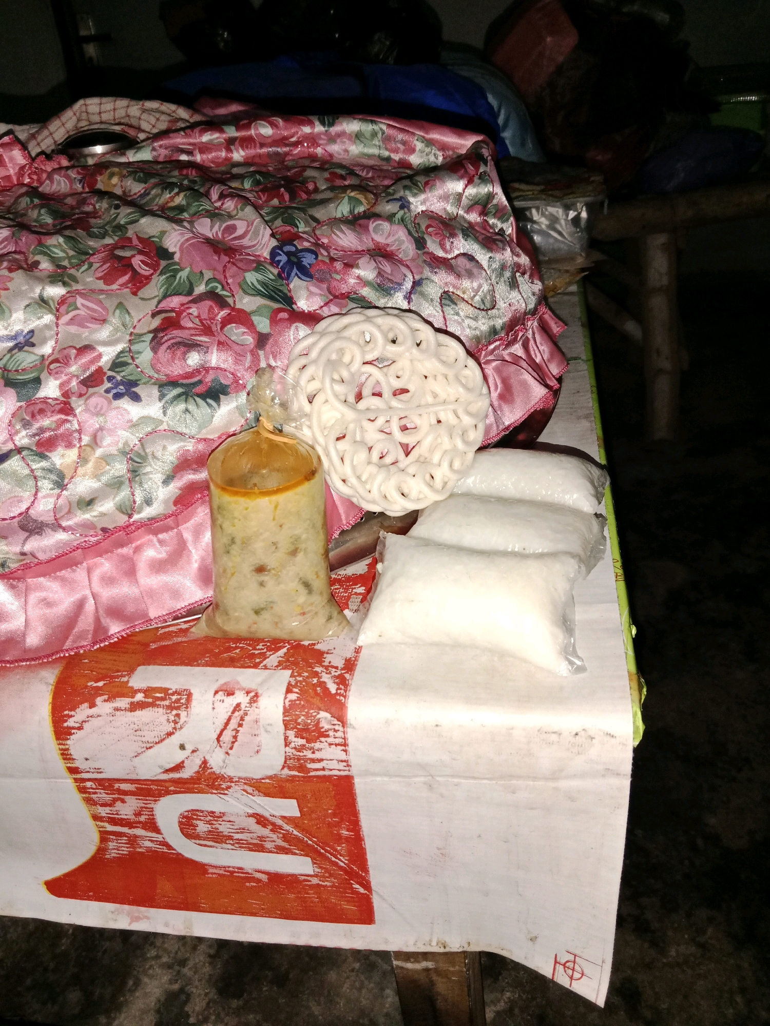 A close-up, slightly blurry, eye-level shot captures a food stall display in Ringinrejo, Indonesia. The foreground is dominated by a white paper tablecloth with large, distressed orange lettering and a red symbol in the bottom right corner. On the tablecloth, three individually packaged white rice cakes, wrapped in clear plastic, are arranged horizontally. To their left, a tall, clear plastic bag contains a yellowish, textured food, possibly rice porridge or a similar dish, sealed at the top with an orange rubber band. Behind these food items, a white, circular, intricately patterned cracker sits. A satin fabric with a pink floral pattern and a ruffled pink trim is draped over the scene, partially obscuring the background. The stall appears to be set up on a wooden table. The lighting is dim, suggesting an evening or indoor setting.