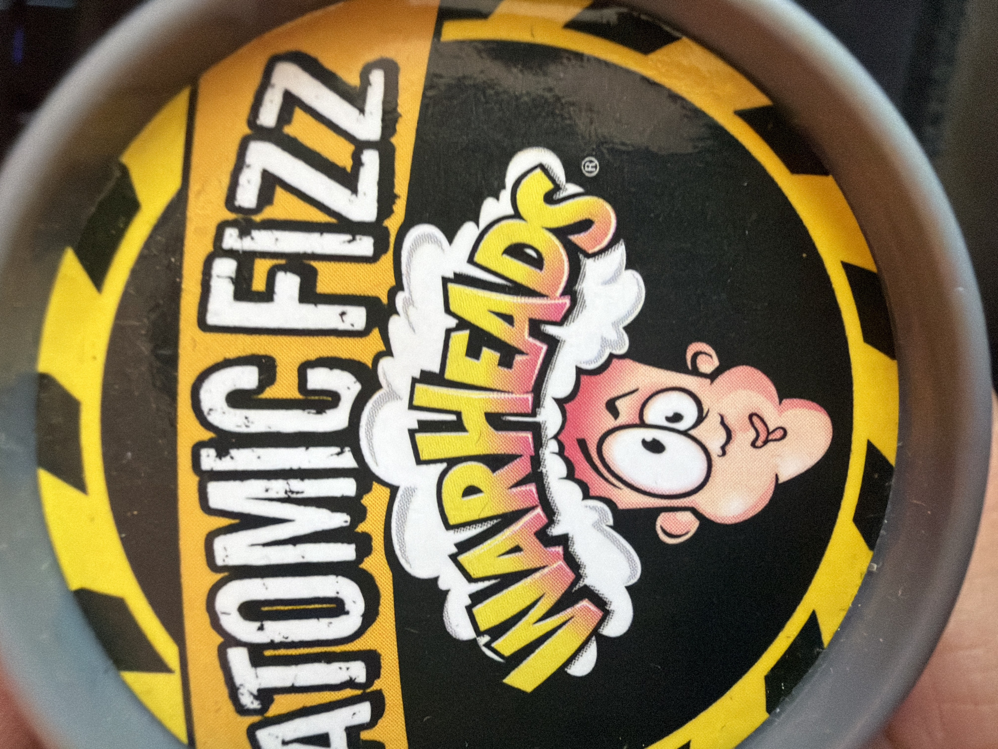 The image shows the top of a container of "Warheads Atomic Fizz" candy. The container is round and made of gray plastic. The label is circular, with a yellow and black hazard stripe border. The words "ATOMIC FIZZ" are stacked vertically in white letters on the left side. A cartoon character with a reddish-pink head is positioned in the center of the label. The word "WARHEADS" is displayed in yellow and orange letters with white clouds around it. There is no indication of the location being in Sofia, Bulgaria.