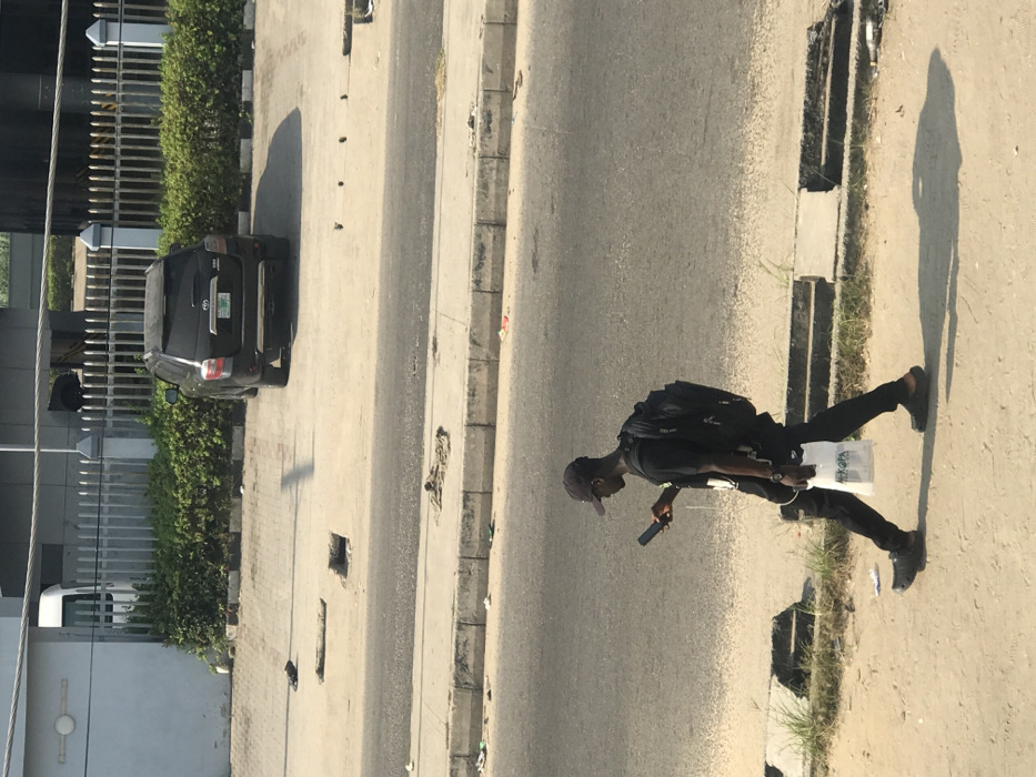 The image displays a daytime street scene in the city of Ado, Nigeria, under bright sunlight, evidenced by prominent shadows.
In the lower foreground, a dark-skinned male adult walks from right to left on a dirt path. He is dressed in a dark baseball cap, dark sunglasses, a dark t-shirt, dark trousers, and dark slip-on shoes. He holds a black smartphone in his right hand, looking down at the screen. A black backpack is worn on his back, and his left hand carries a white plastic shopping bag with "EKOPA" printed on it. White earphones or a cable hang from his neck.
Behind the pedestrian, an asphalt road stretches horizontally, bordered by a concrete curb littered with debris. Beyond the road, an elevated, light-colored paved area contains multiple square depressions or potential drainage openings.
In the background, a dark-colored Toyota Highlander SUV is parked facing away from the viewer on a paved surface, exhibiting a green Nigerian license plate. Behind the parked SUV and the paved area, a grey metal fence with vertical pickets and horizontal rails, topped with security wire, is visible, fronted by green bushes. To the left of the fence, a light-colored building and a partial white van are present. To the right, dark cylindrical industrial structures are partially visible behind the fence. A thick grey cable runs horizontally across the top section of the frame.