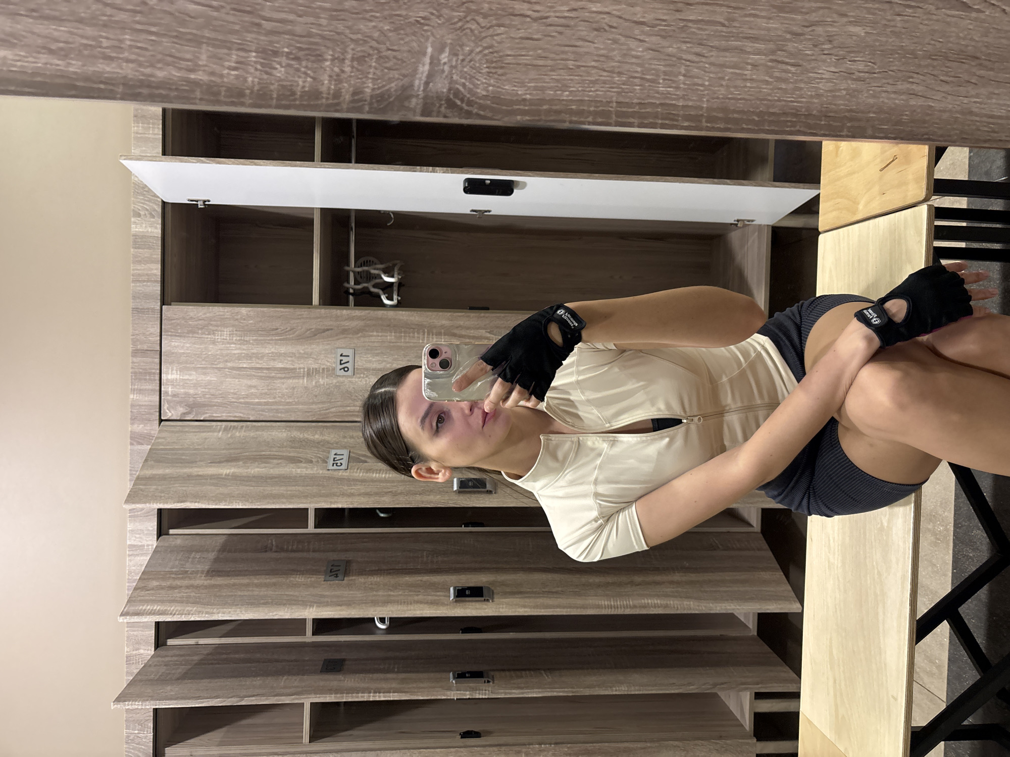 A young woman is taking a selfie in what appears to be a locker room. The background consists of rows of wooden lockers with numbers. She is wearing workout clothes: a light-colored jacket and dark shorts, and gloves. She is holding a phone in her left hand to take the photo. The location may be in Krasnoyarsk, Russia, but there is no direct indication from the image itself.