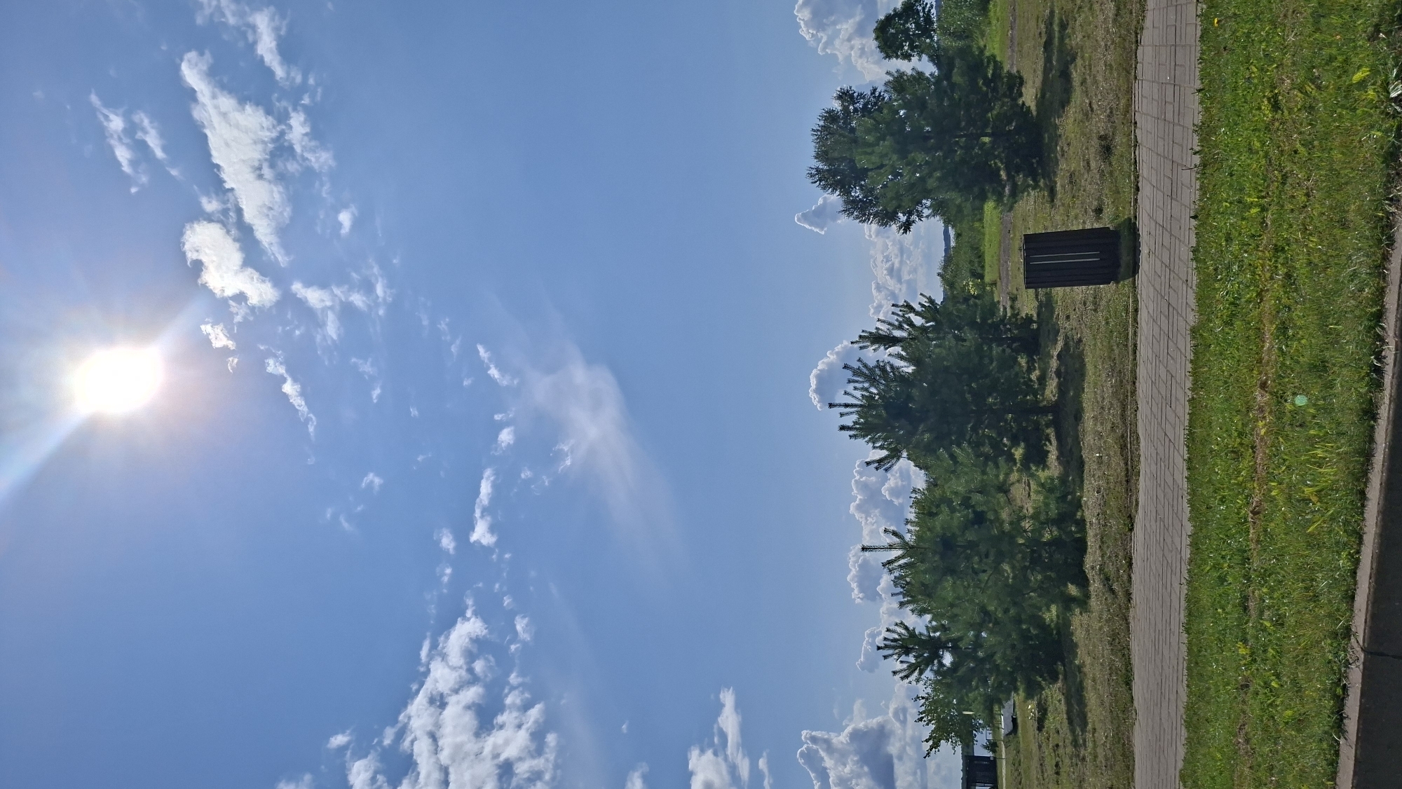 The image depicts a bright, sunny day with a clear blue sky and scattered white clouds. The sun is high in the sky, casting a bright glare. In the foreground, there is a paved walkway made of interlocking gray stones, bordered by a strip of green grass. Beyond the walkway is a grassy area with several small evergreen trees and a larger, fuller green tree to the right. In the background, there are more trees and a body of water, possibly a lake, with a small structure and a boat visible near the left edge of the frame. A dark, rectangular trash receptacle stands near the walkway and trees on the right side of the image. The scene appears to be a park or recreational area. There are no people or animals visible in the image. The location is identified as Myadzyel, Belarus.